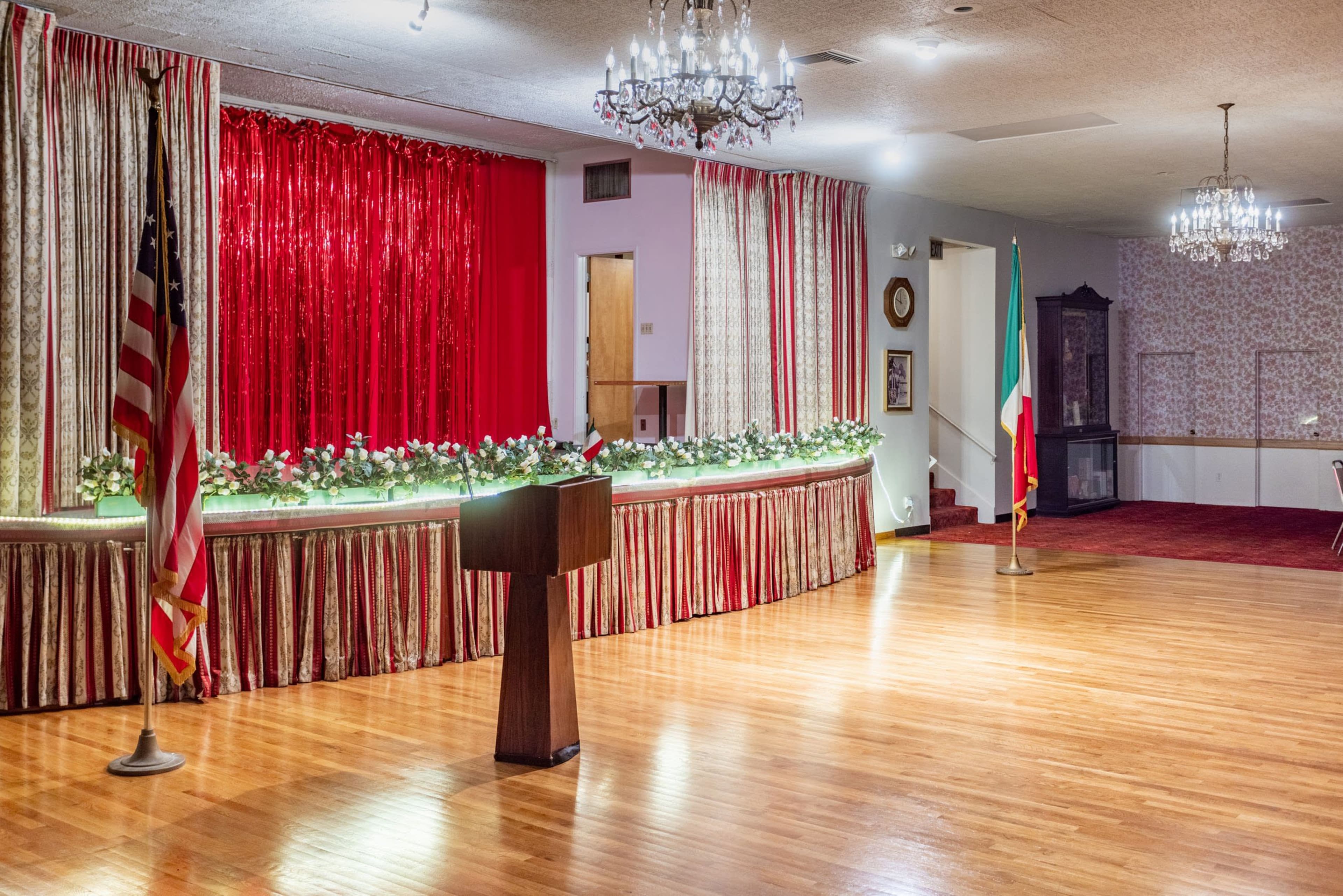 The image shows a decorated event hall with a podium in front of a red and white curtain backdrop, flanked by floral arrangements, an American flag, and an Italian flag.