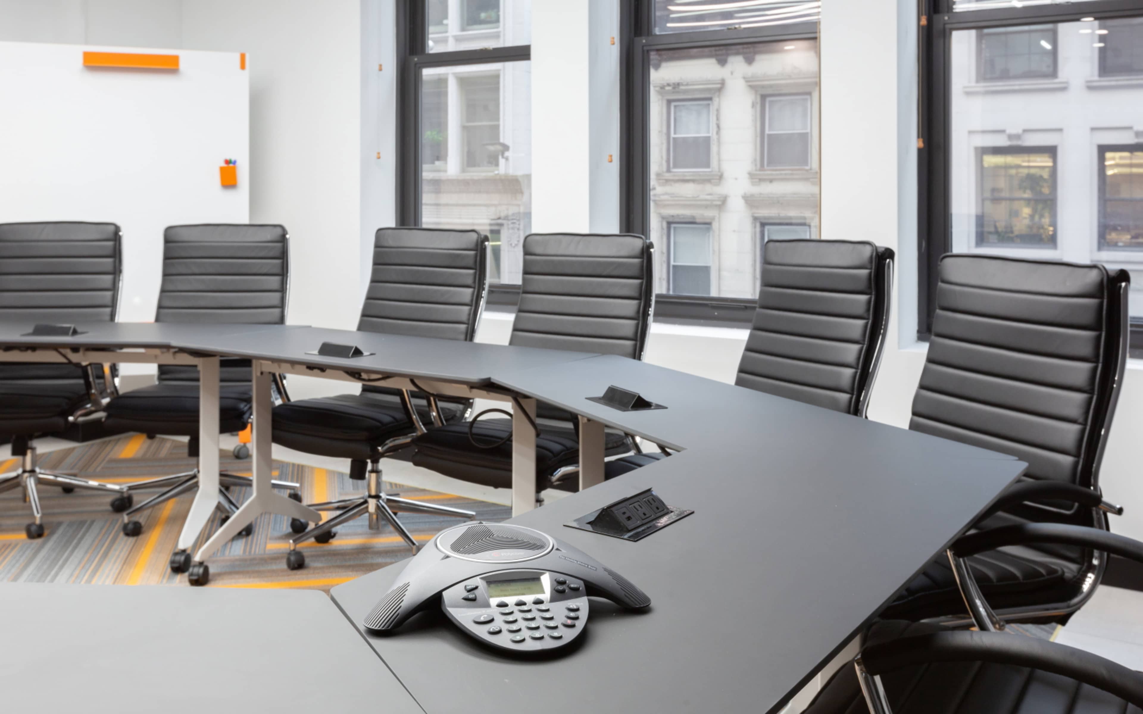 A modern conference room features a curved table with several black ergonomic chairs and a conference phone in the center.