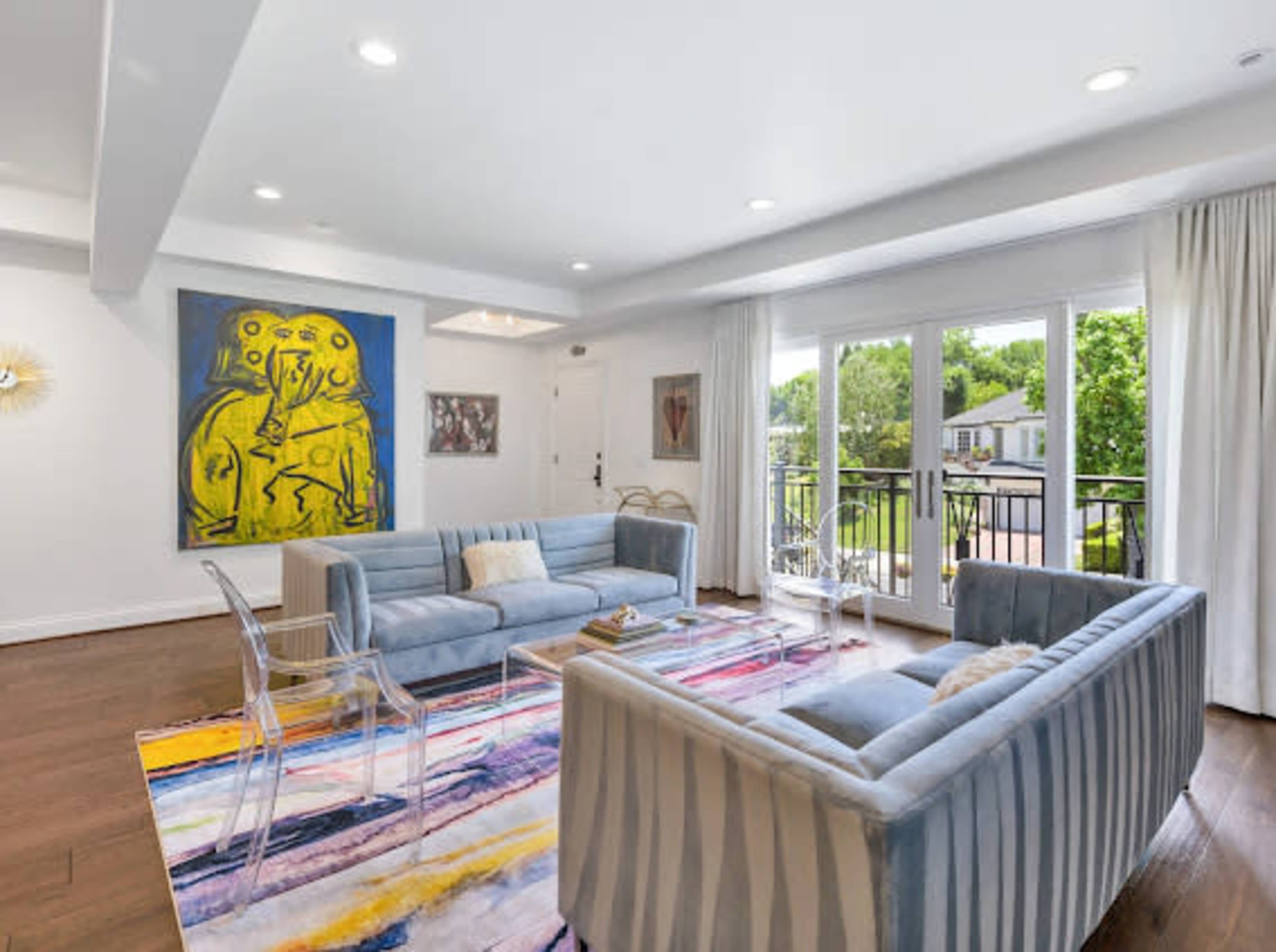A modern living room features two striped sofas, a colorful area rug, a large painting on the wall, and sliding glass doors leading to a balcony.