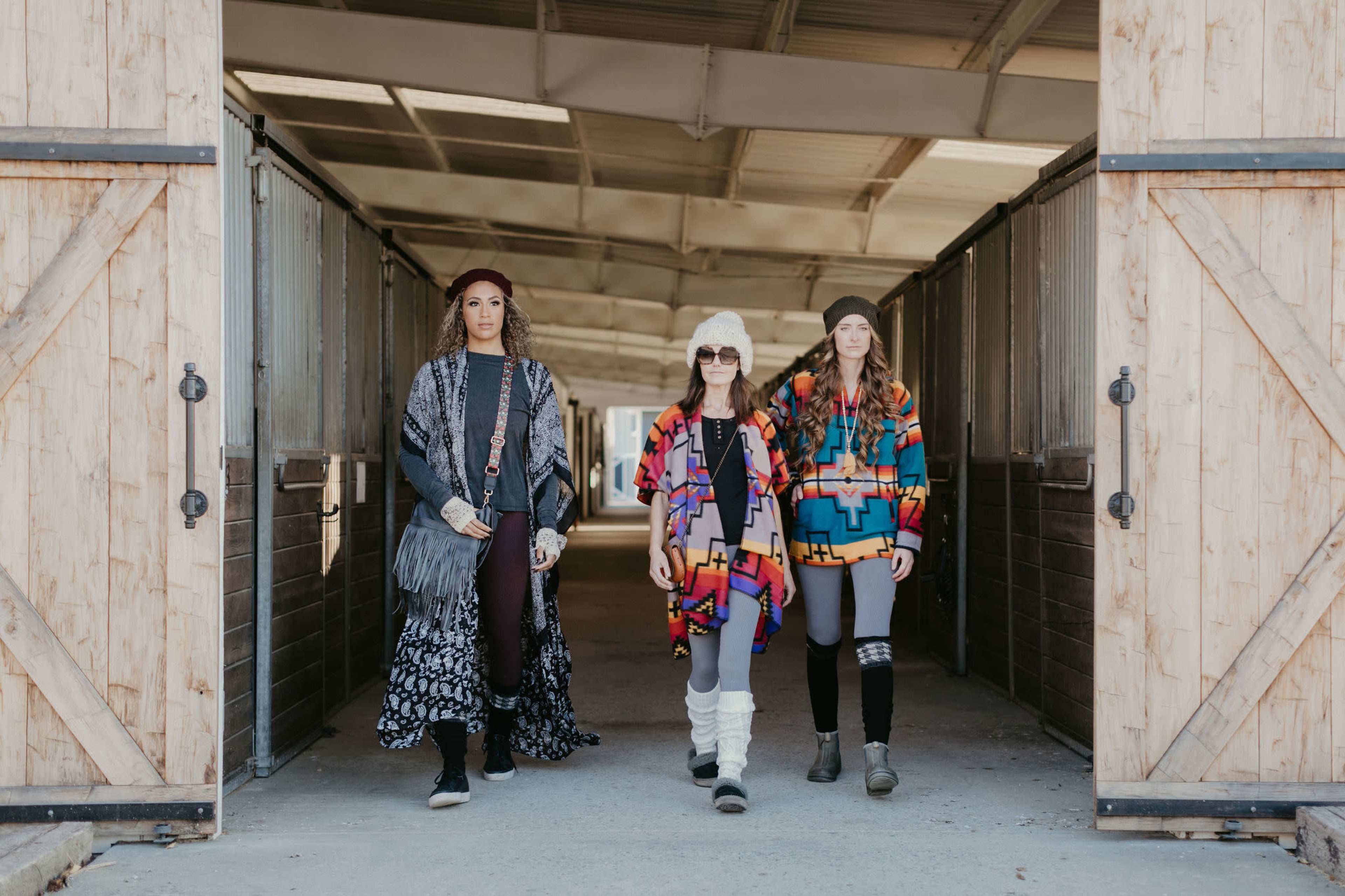 Three women are walking down a barn aisle wearing colorful, patterned outfits and accessories.