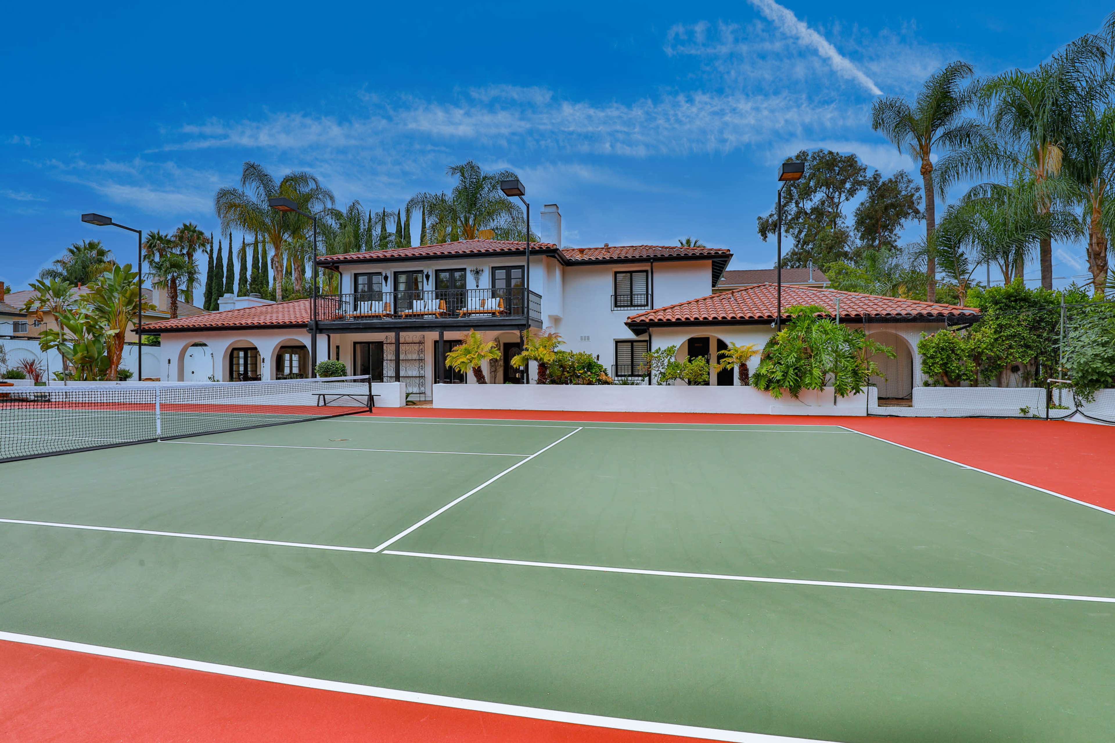 A tennis court in front of a two-story house surrounded by palm trees and greenery.