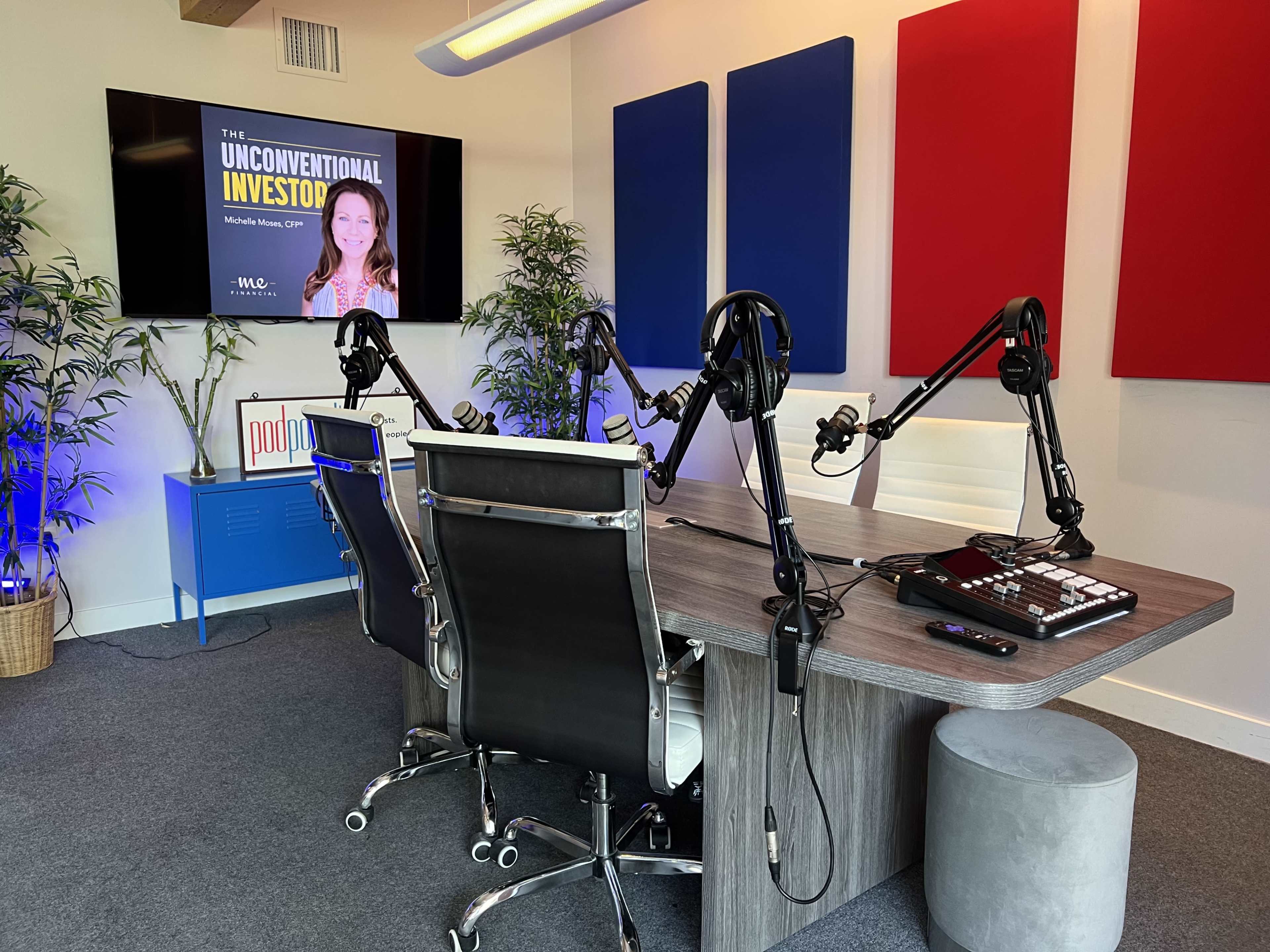 A modern podcast studio features a table with four chairs, mounted microphones, and a screen displaying "The Unconventional Investor" with a backdrop of blue and red panels.