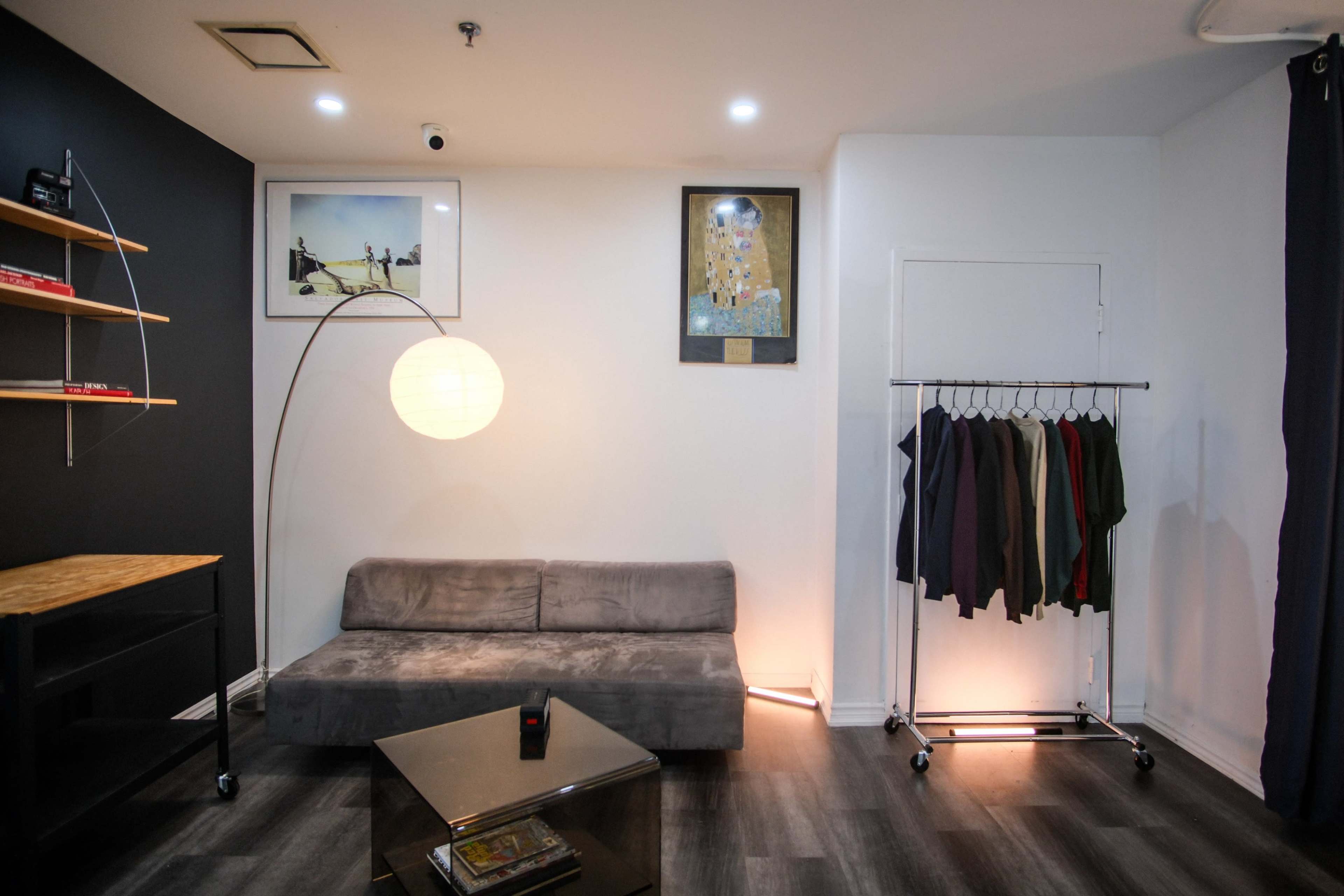 A small, modern room with a gray sofa, a clothing rack with hanging garments, a circular lamp, and various wall art.