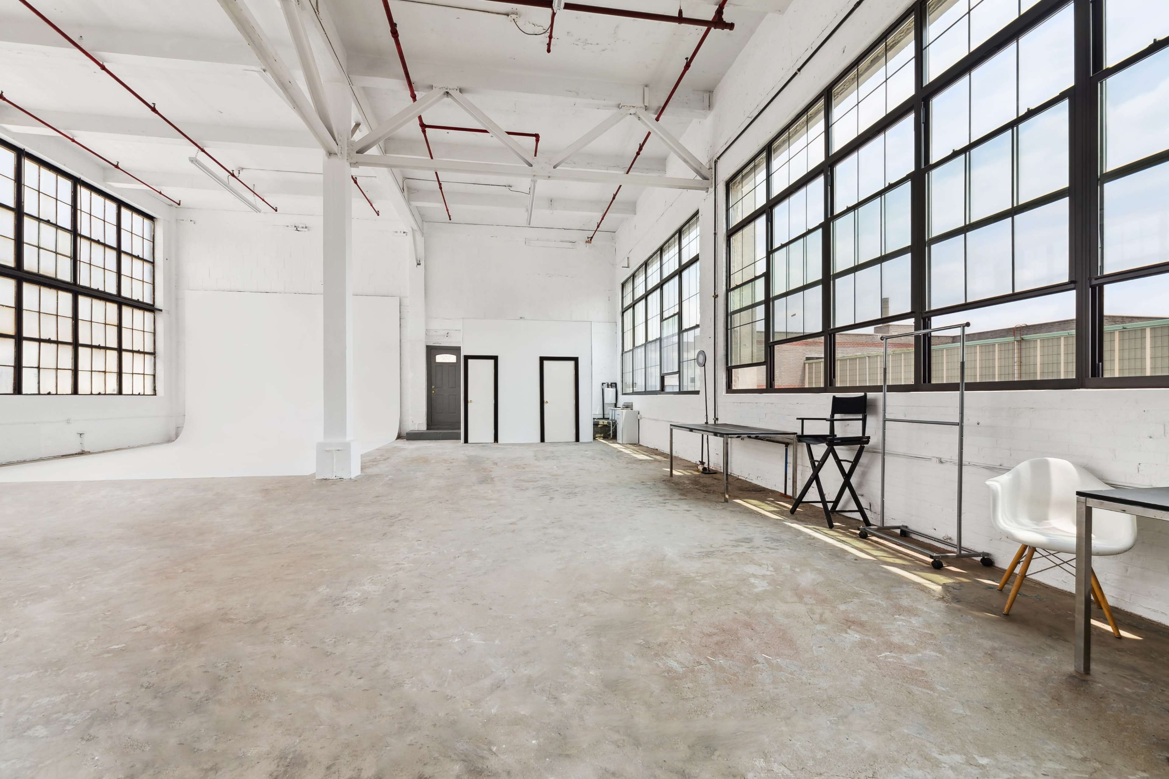 A spacious, well-lit studio with large windows, concrete flooring, and minimal furnishings.
