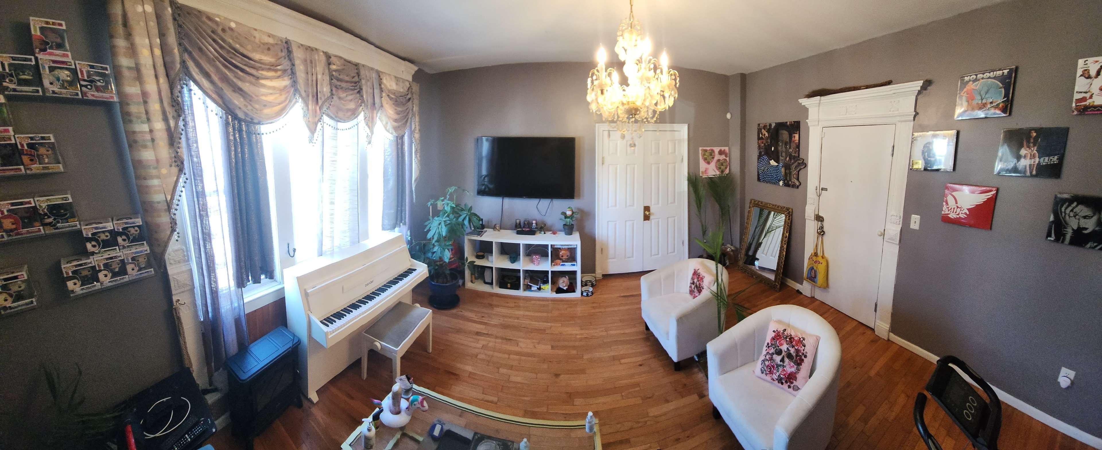 A well-furnished living room with a piano, two white chairs, a television mounted on the wall, and decorative artwork arranged on the walls.