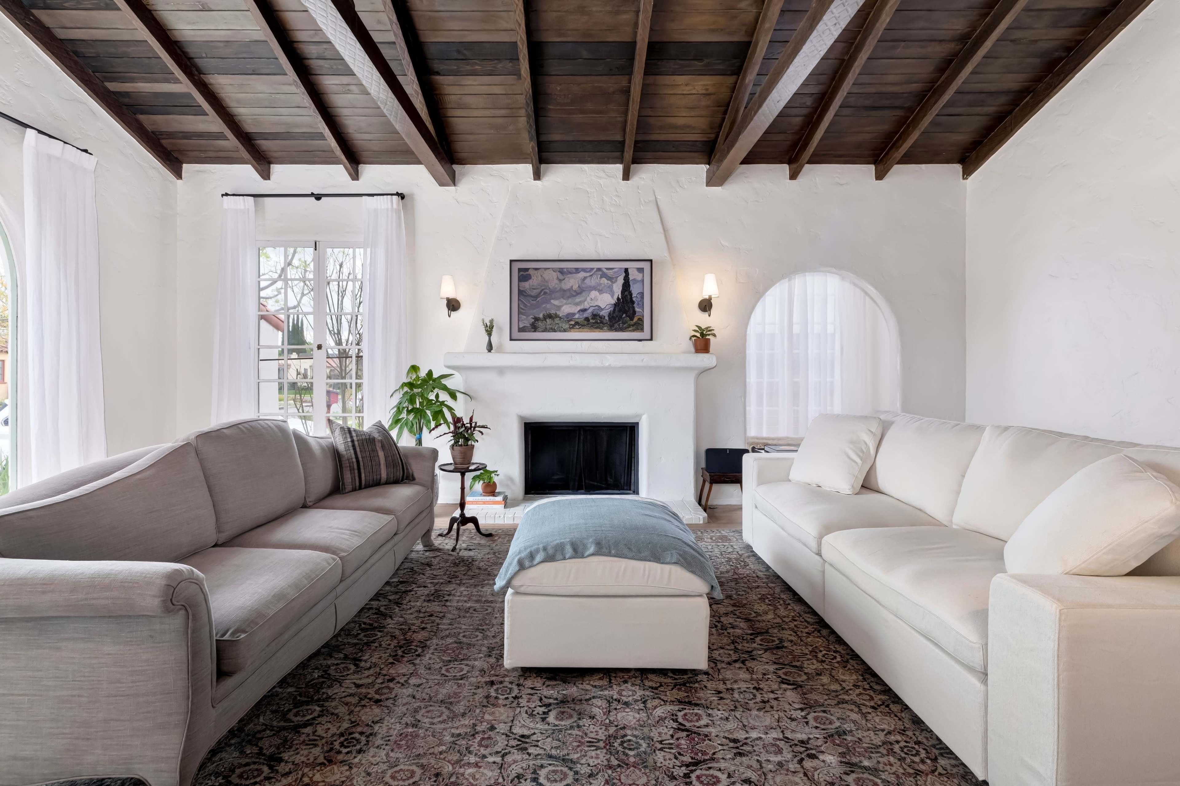 A bright living room features two sofas, an ottoman, a fireplace, and a decorative plant under wooden beams.