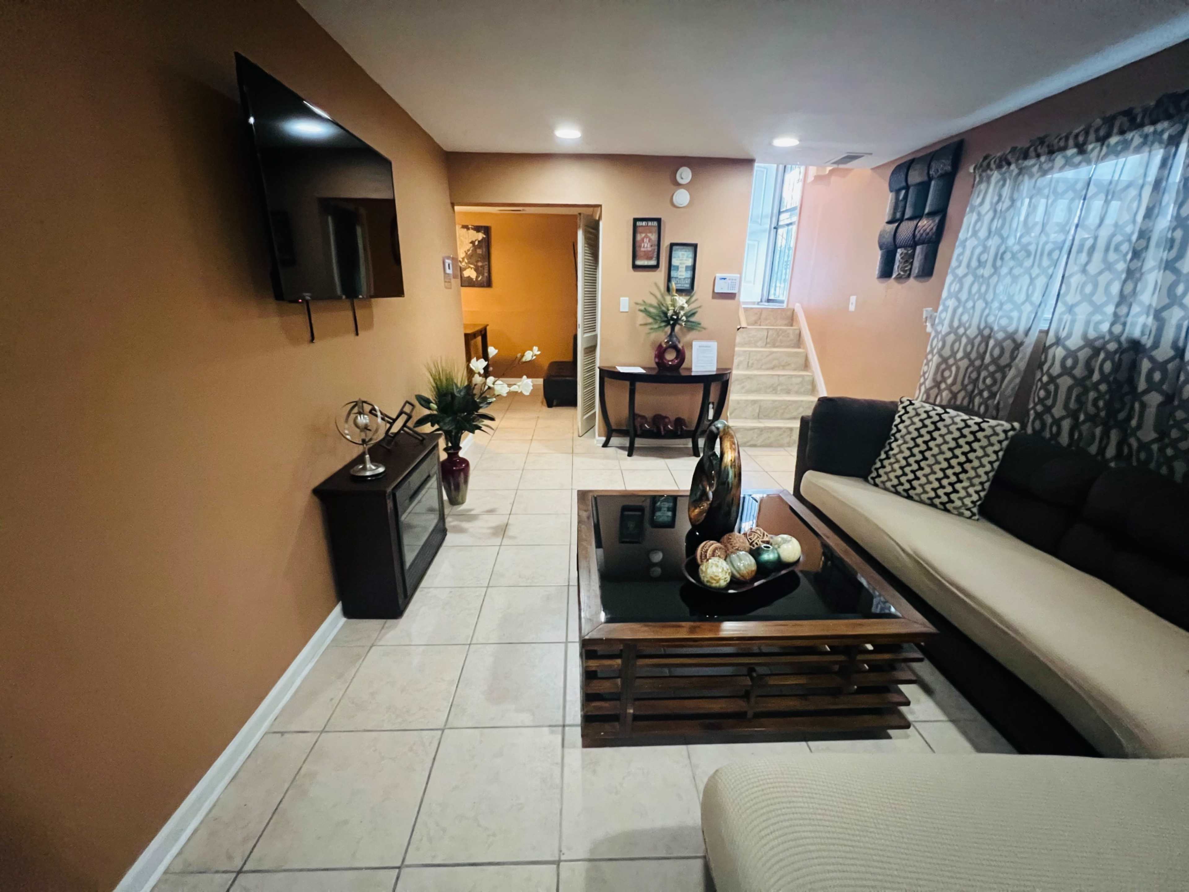 A living room with a flat-screen TV mounted on the wall, a coffee table in the center, and seating arrangements on a tiled floor.