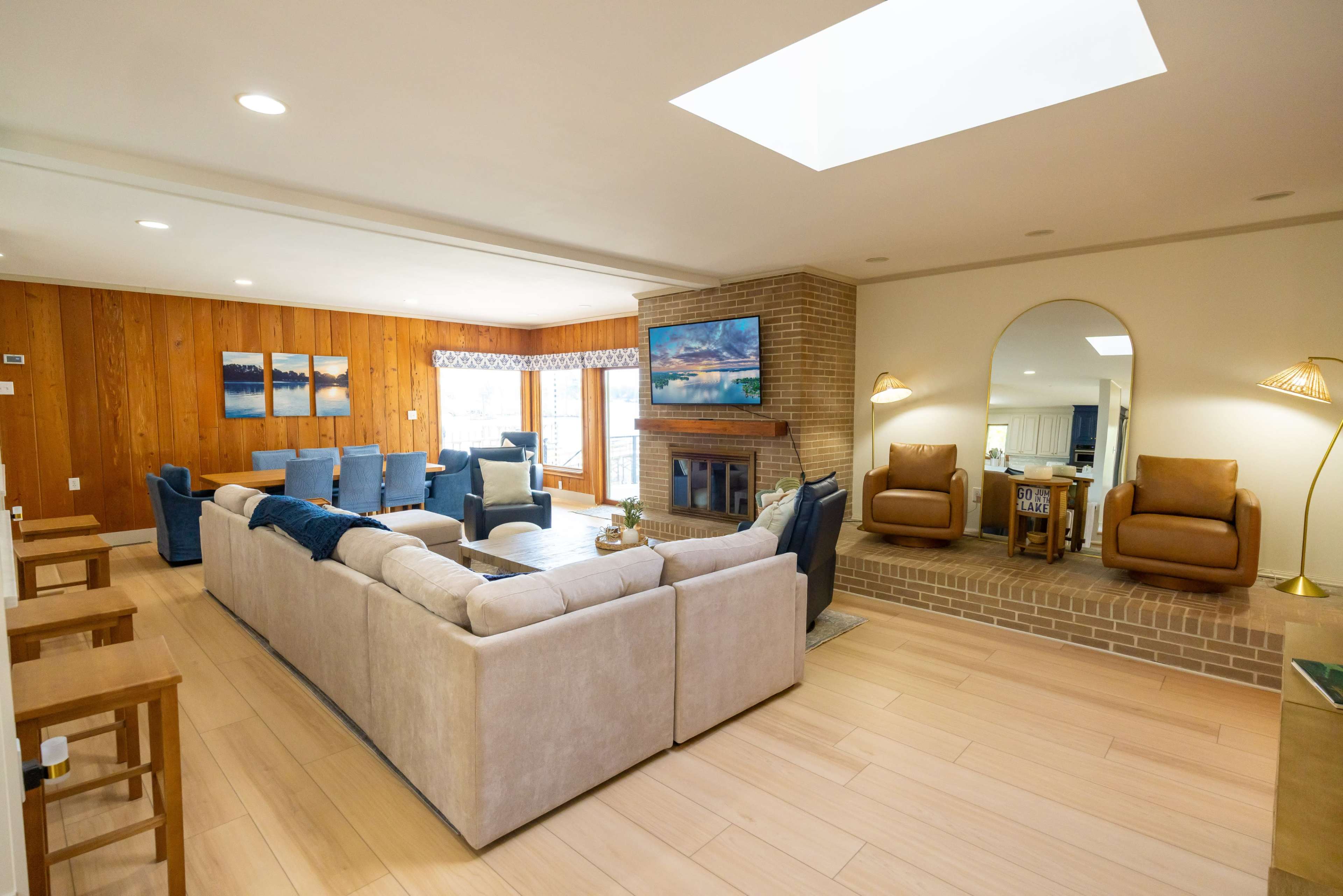 The image shows a spacious living room with a large sectional sofa, a fireplace, and a skylight, featuring wooden paneling and a view of the outdoors through large windows.