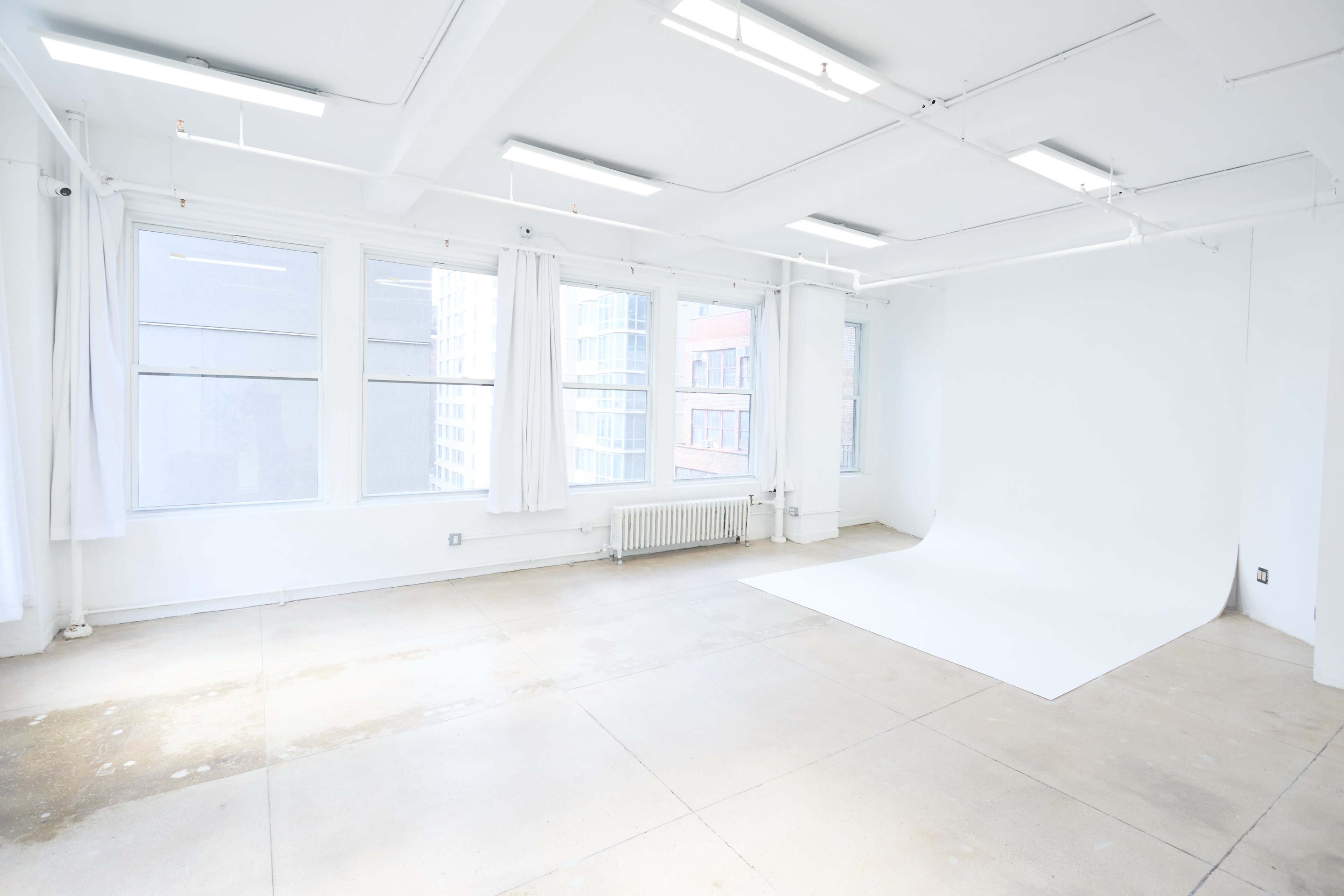 The image shows a bright, empty studio space with large windows, white walls, and a clean backdrop.