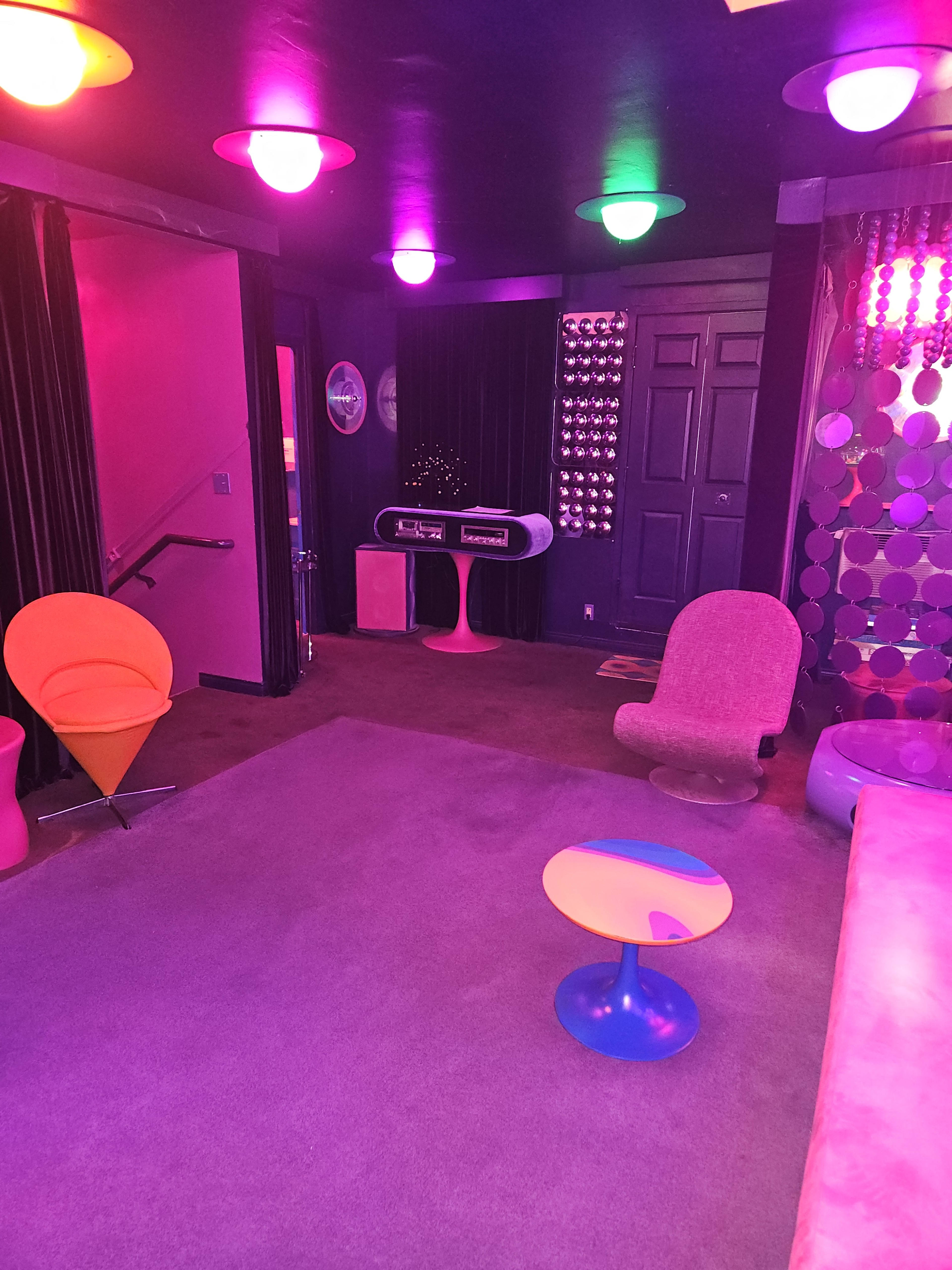 The image shows a room decorated with vibrant pink and purple lighting, featuring modern furniture like colorful chairs and circular tables.