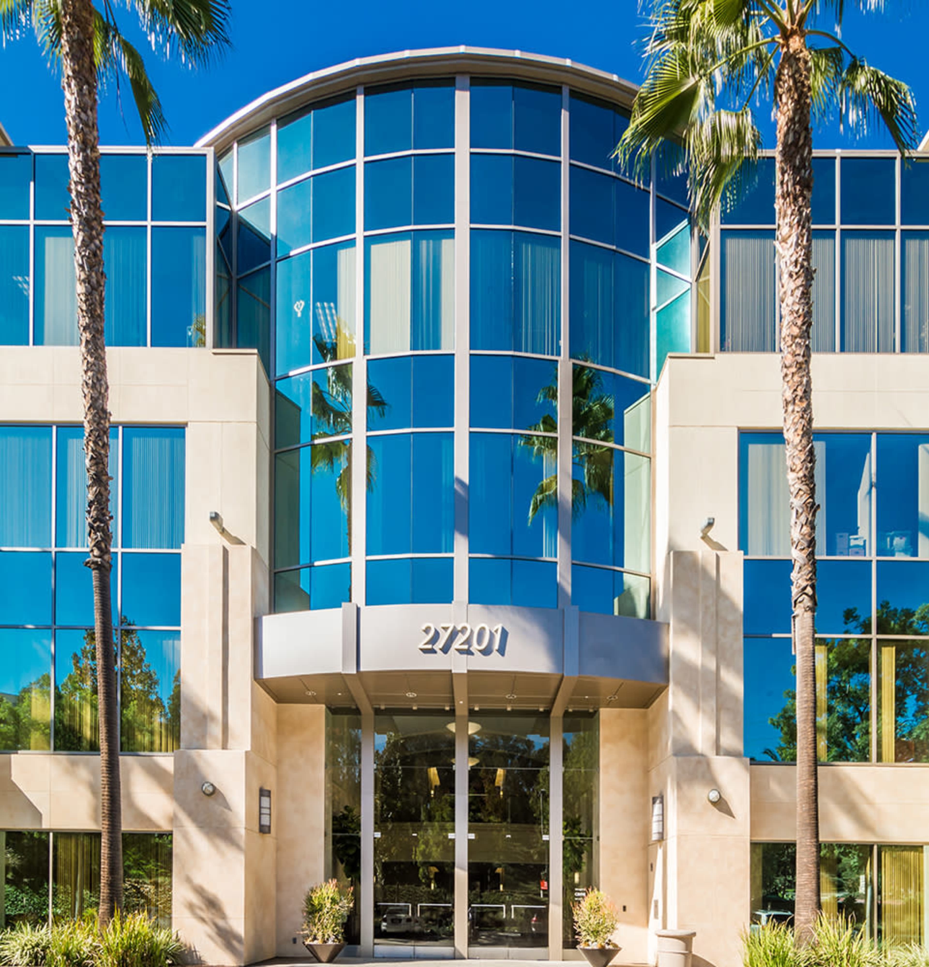 A modern glass-fronted building with a prominent entrance and palm trees in front.