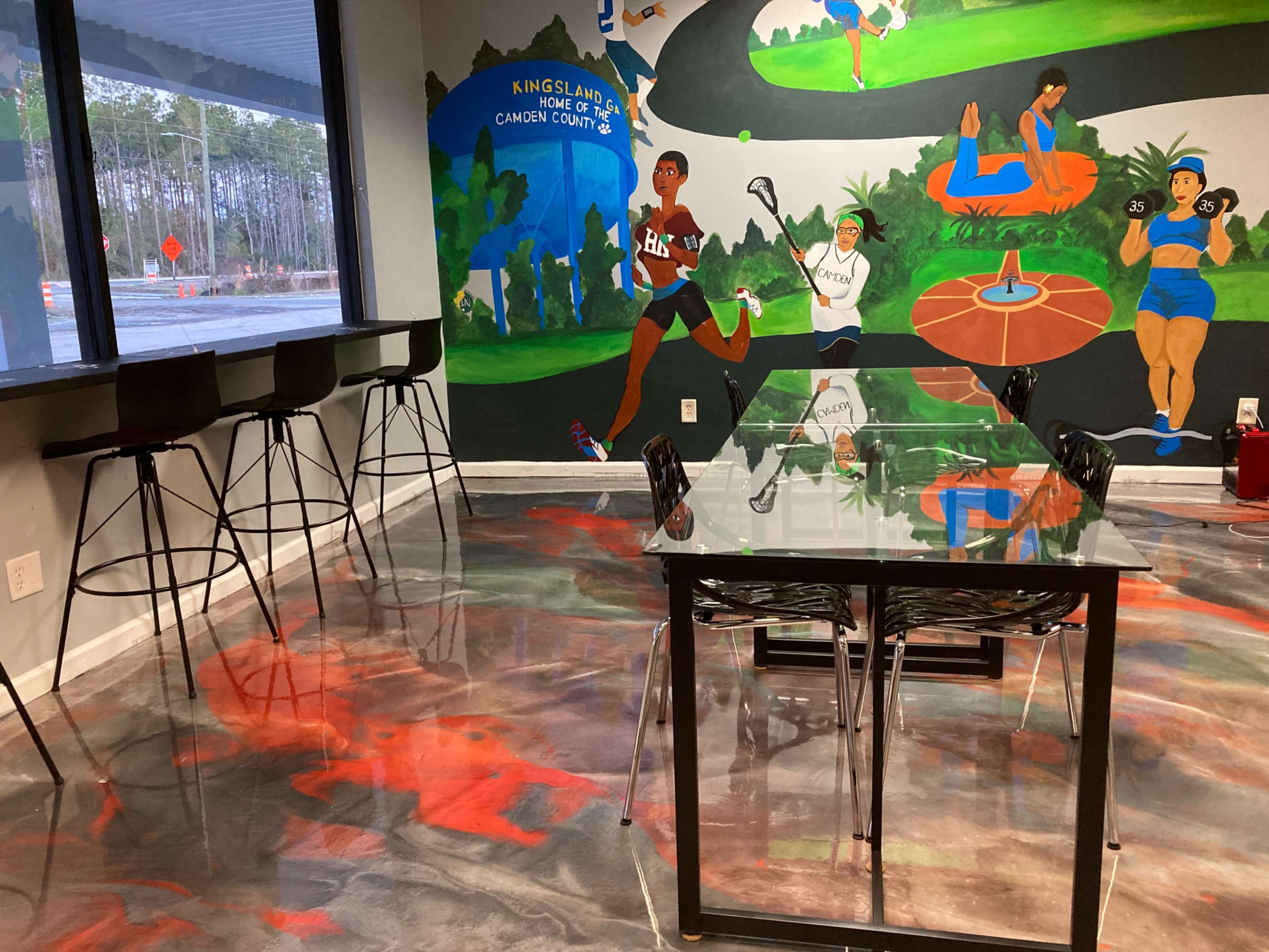 A modern meeting room features a glass table and black chairs, with a colorful mural depicting various sports activities on the wall.