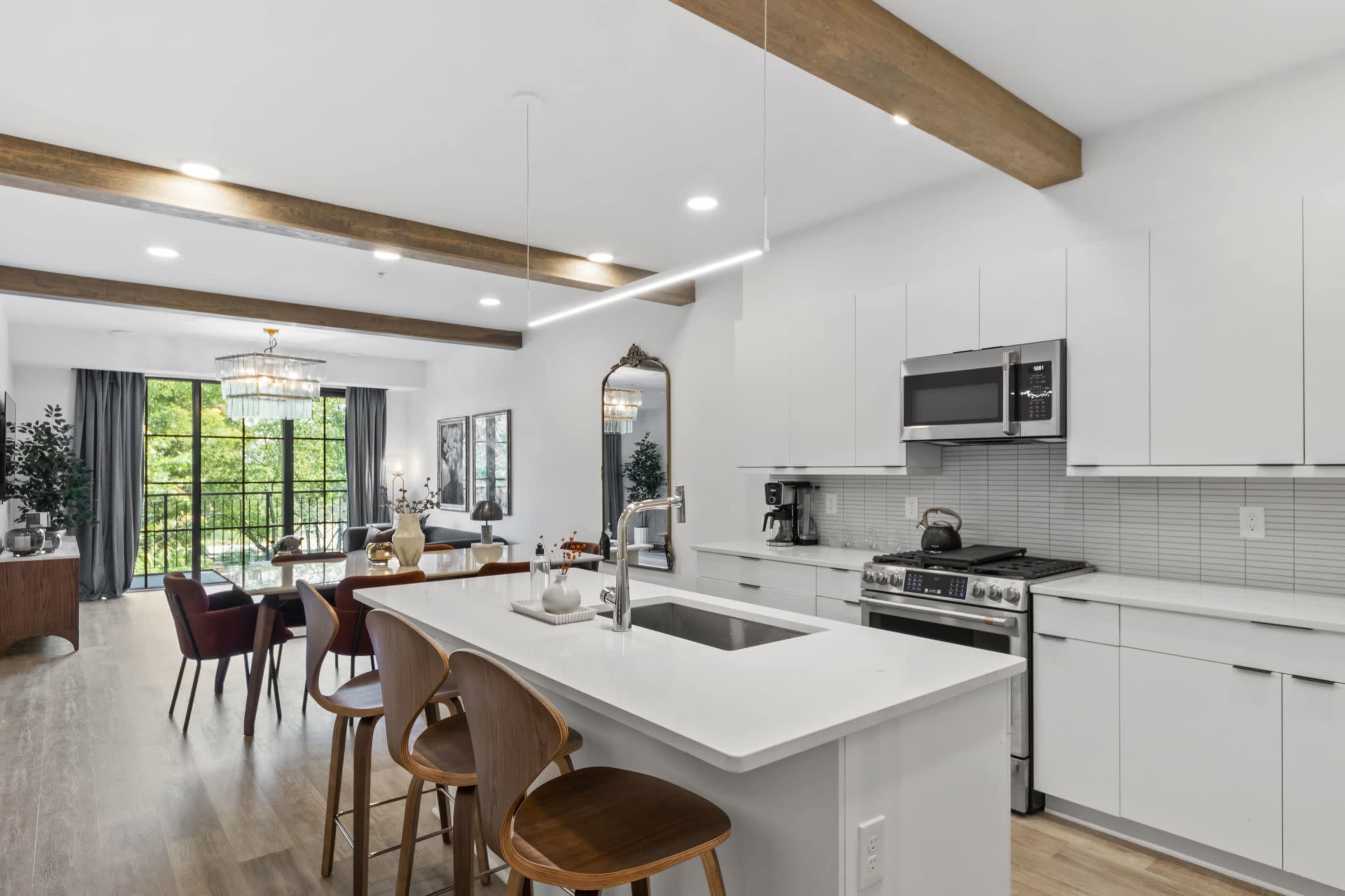 The image shows a modern open-concept kitchen and living area with wooden beams, white cabinetry, and a large window allowing natural light to fill the space.