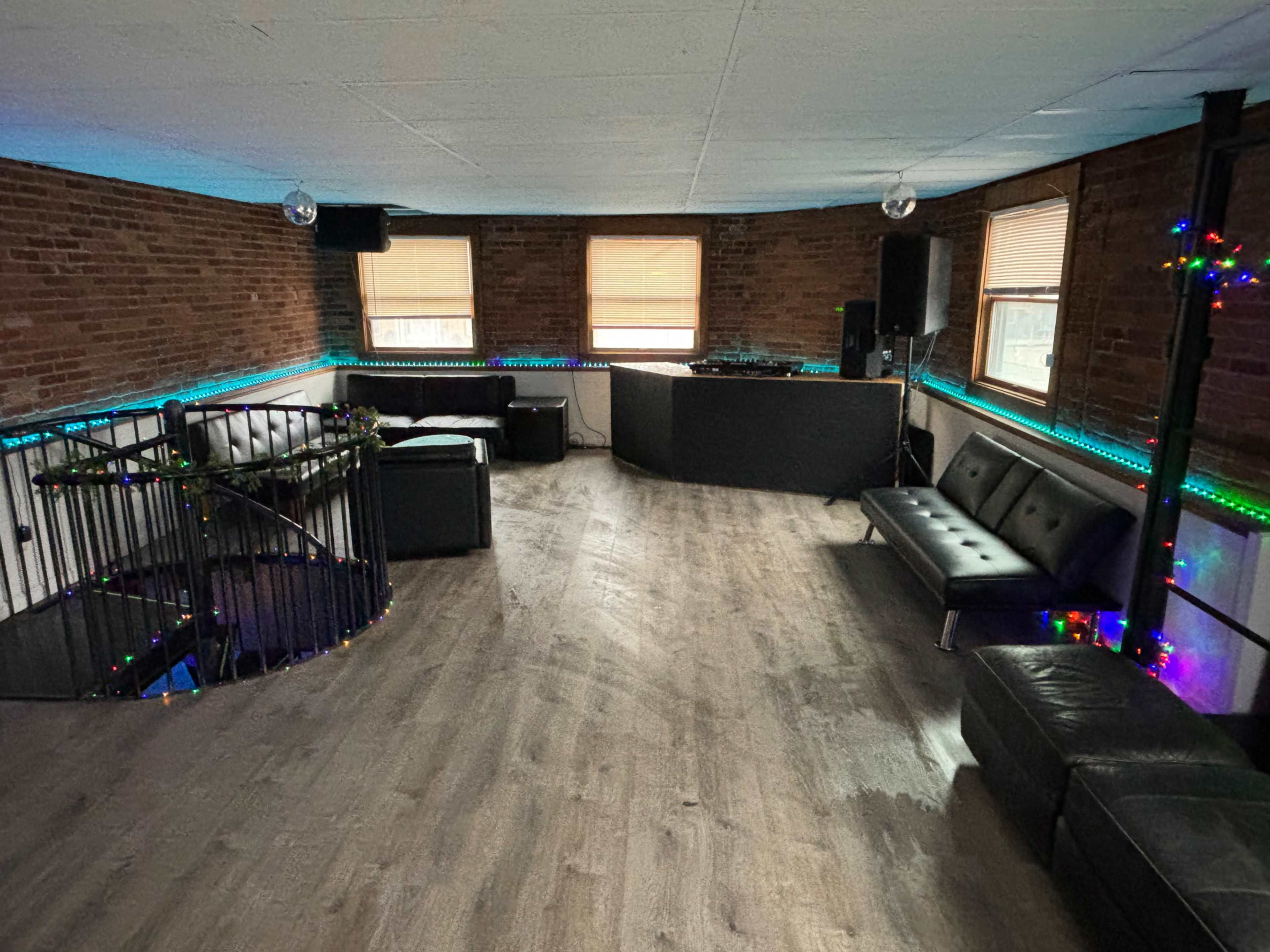 A sparsely furnished room with exposed brick walls, a DJ setup, a spiral staircase, and colored lights along the perimeter.