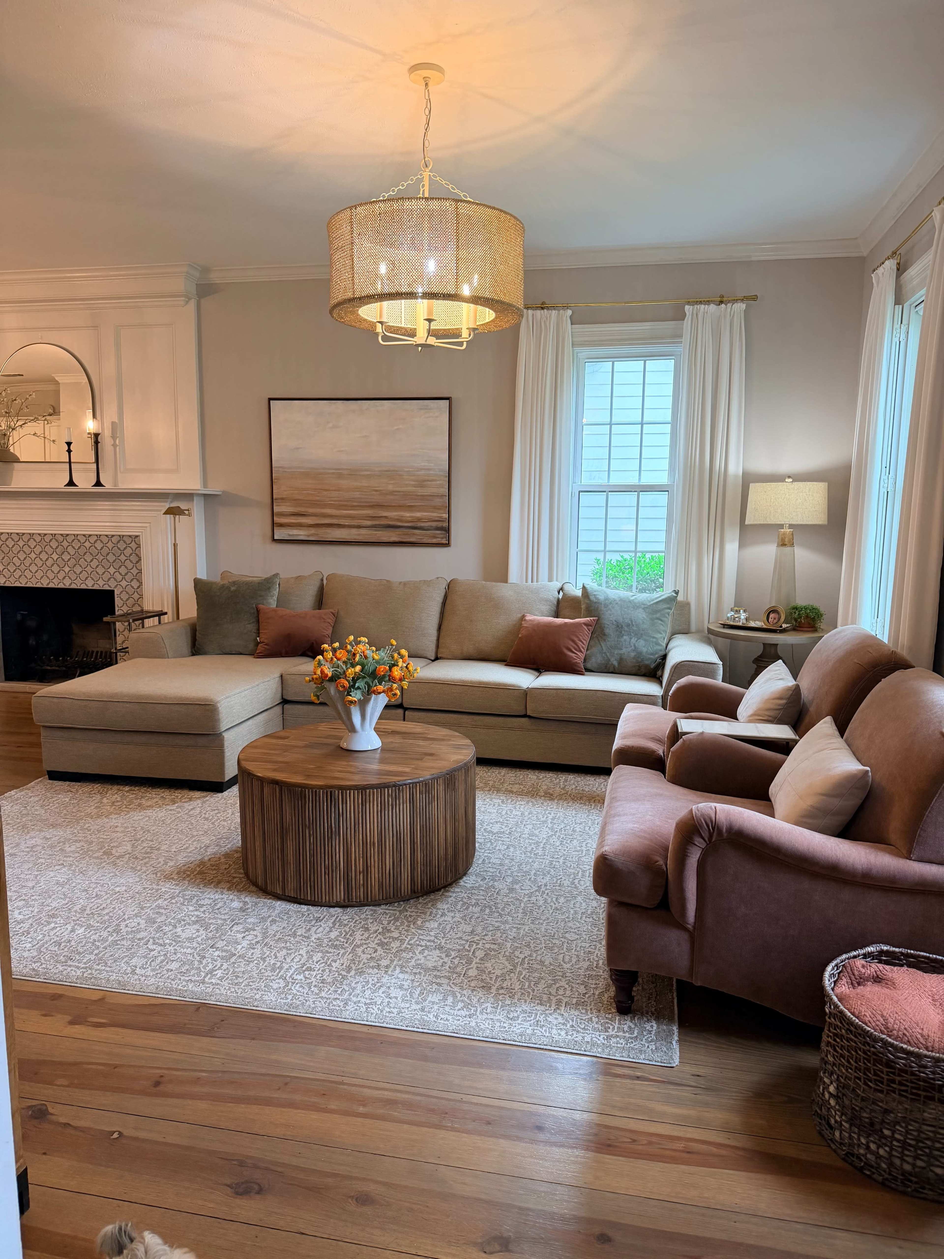 The image shows a cozy living room with a sectional sofa, a round coffee table, and a large window dressed with white curtains.