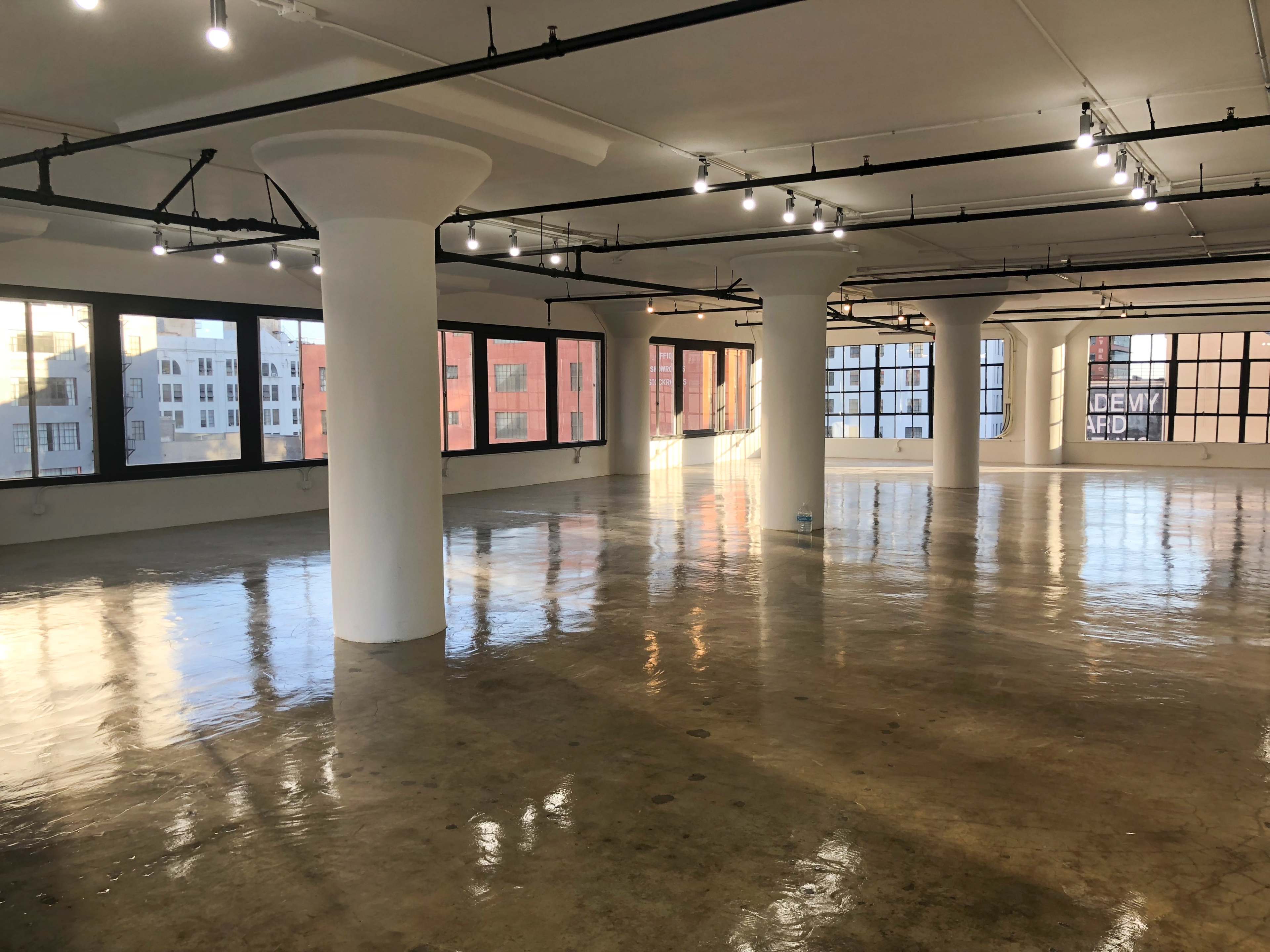 The image shows a spacious, empty room with polished concrete floors and large windows along the walls, illuminated by overhead lights.