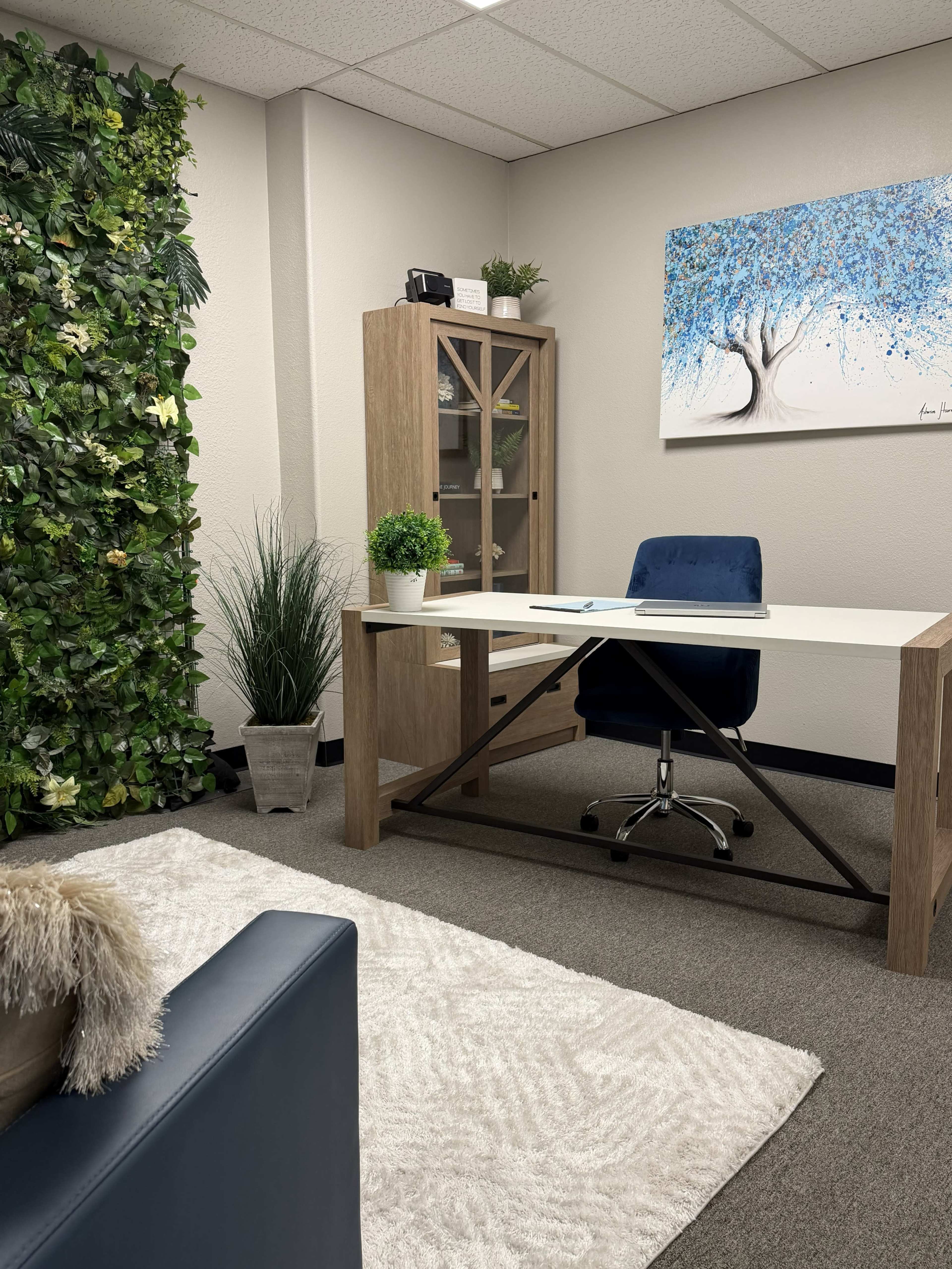 The image shows a modern office space featuring a desk, a blue chair, a cabinet, a rug, and a wall adorned with artificial foliage and artwork.