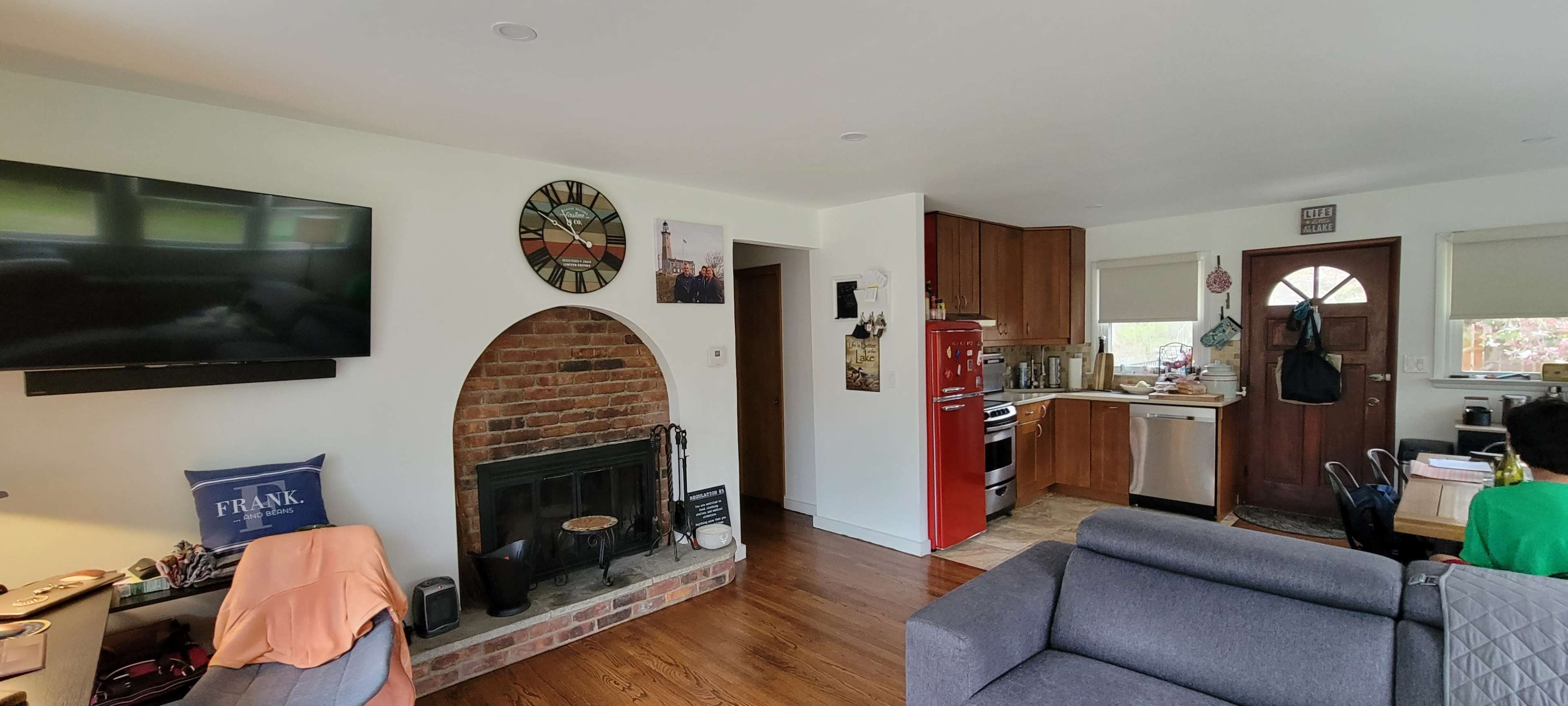 A living room with a brick fireplace, a television on the wall, a red refrigerator in an adjoining kitchen area, and a doorway leading outside.