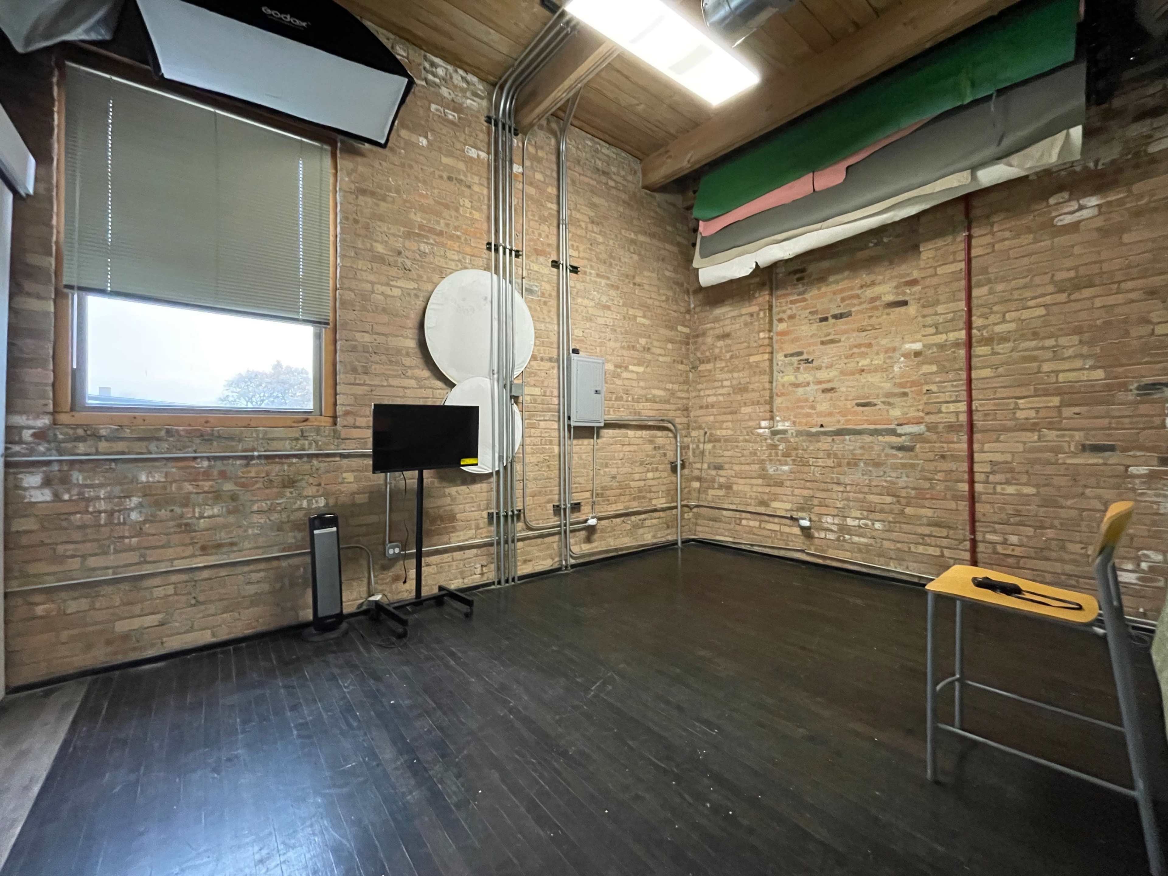 The image shows an empty room with exposed brick walls, a wooden floor, and some visible electrical equipment, including a monitor and chair in the corner.