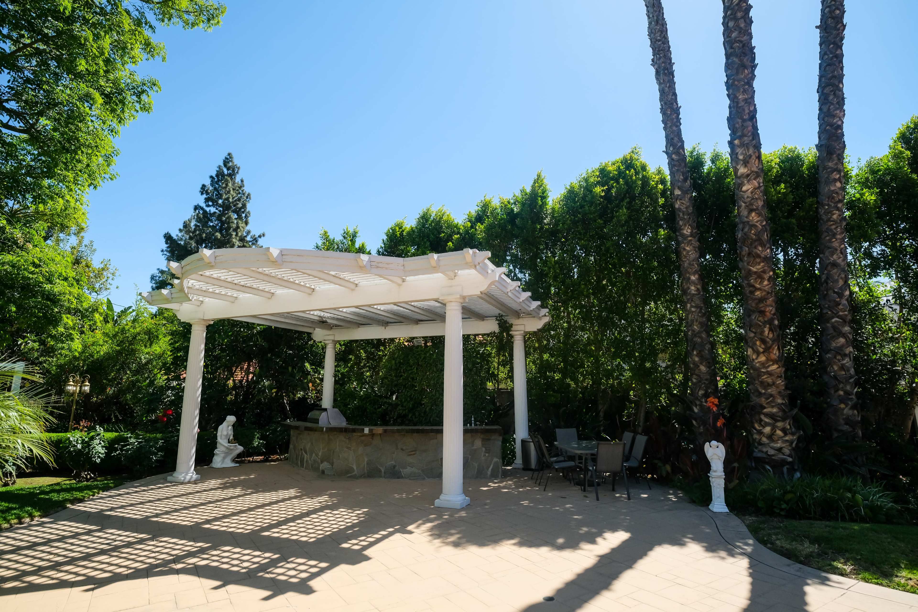 A white pergola with columns provides shade over a stone bar area surrounded by lush greenery and tall palm trees.