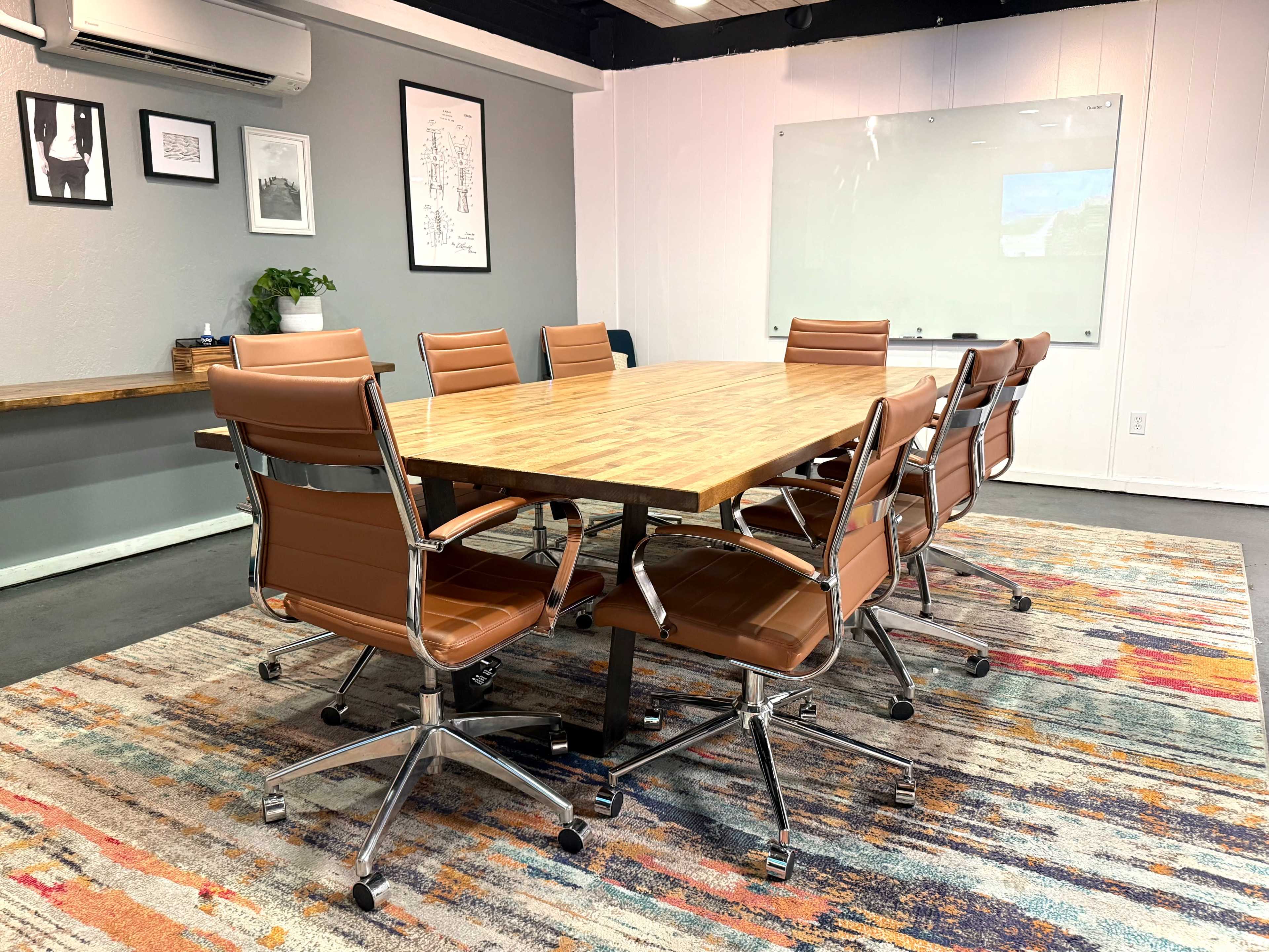 The image shows a modern conference room with a large wooden table surrounded by eight brown leather chairs, a colorful rug, and a whiteboard on the wall.