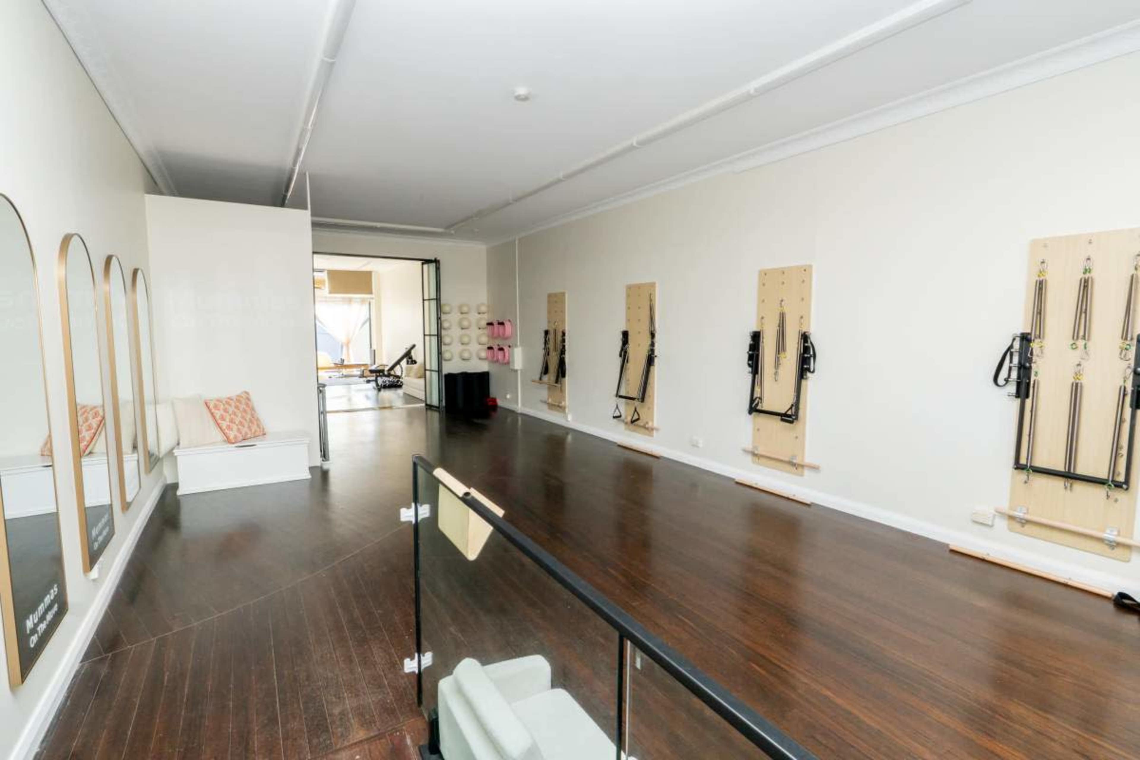 The image shows a spacious, well-lit fitness studio with wooden flooring and wall-mounted exercise equipment arranged neatly along the walls.