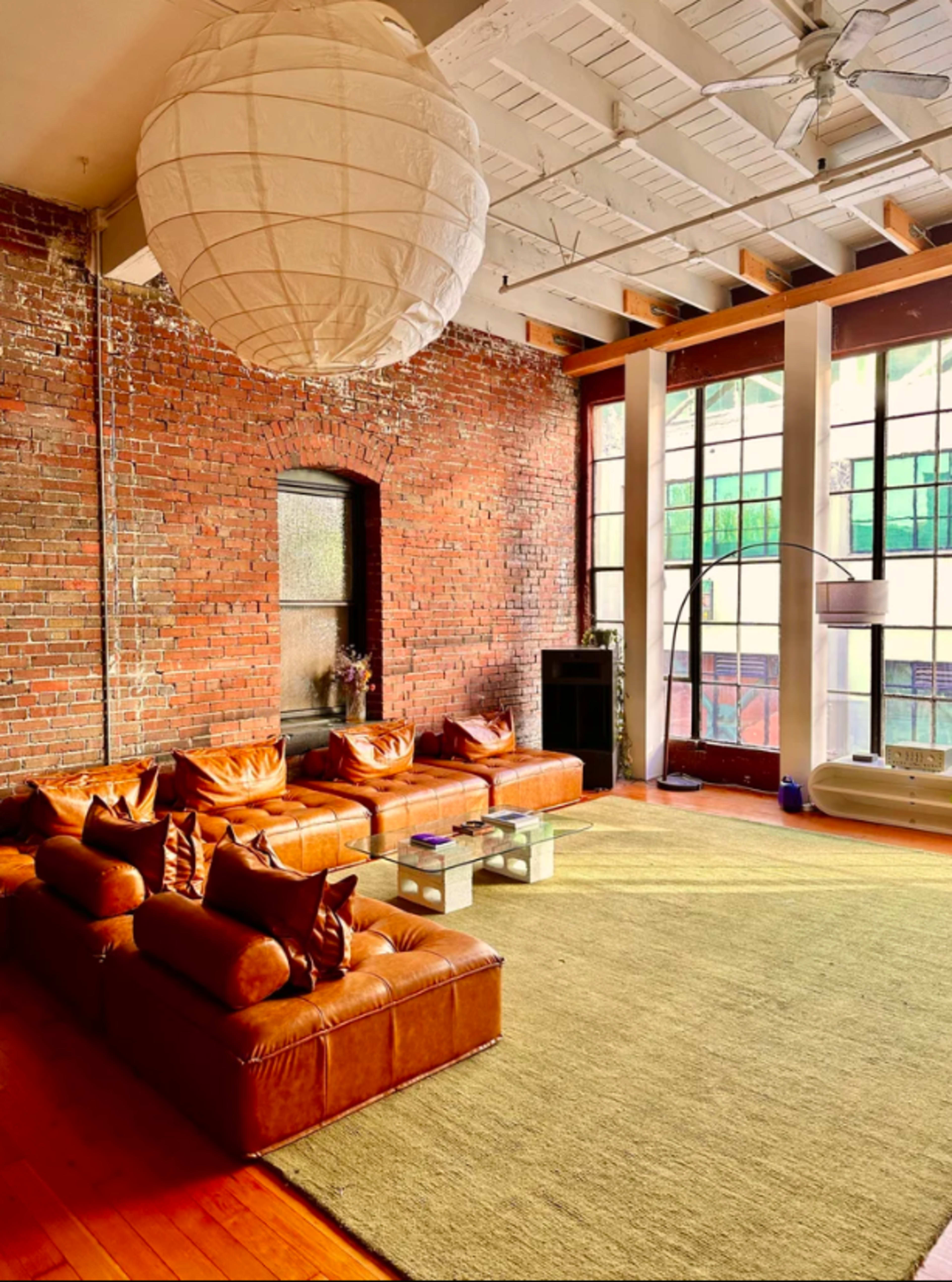 The image shows a spacious living area with exposed brick walls, large windows, and leather seating arranged around a coffee table on a green rug.
