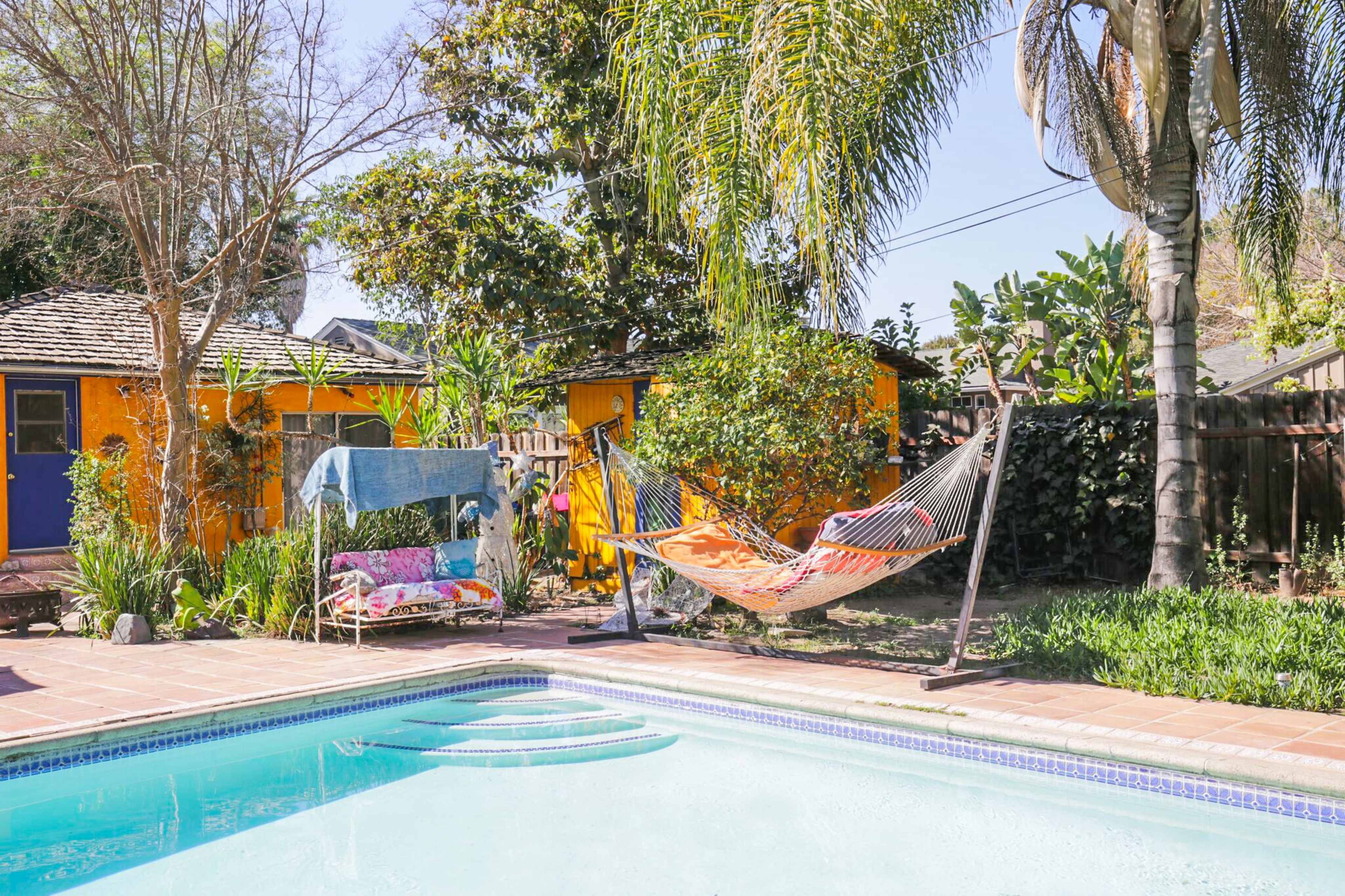 A swimming pool is surrounded by tropical plants, a hammock, and a colorful garden shed in a sunny backyard.