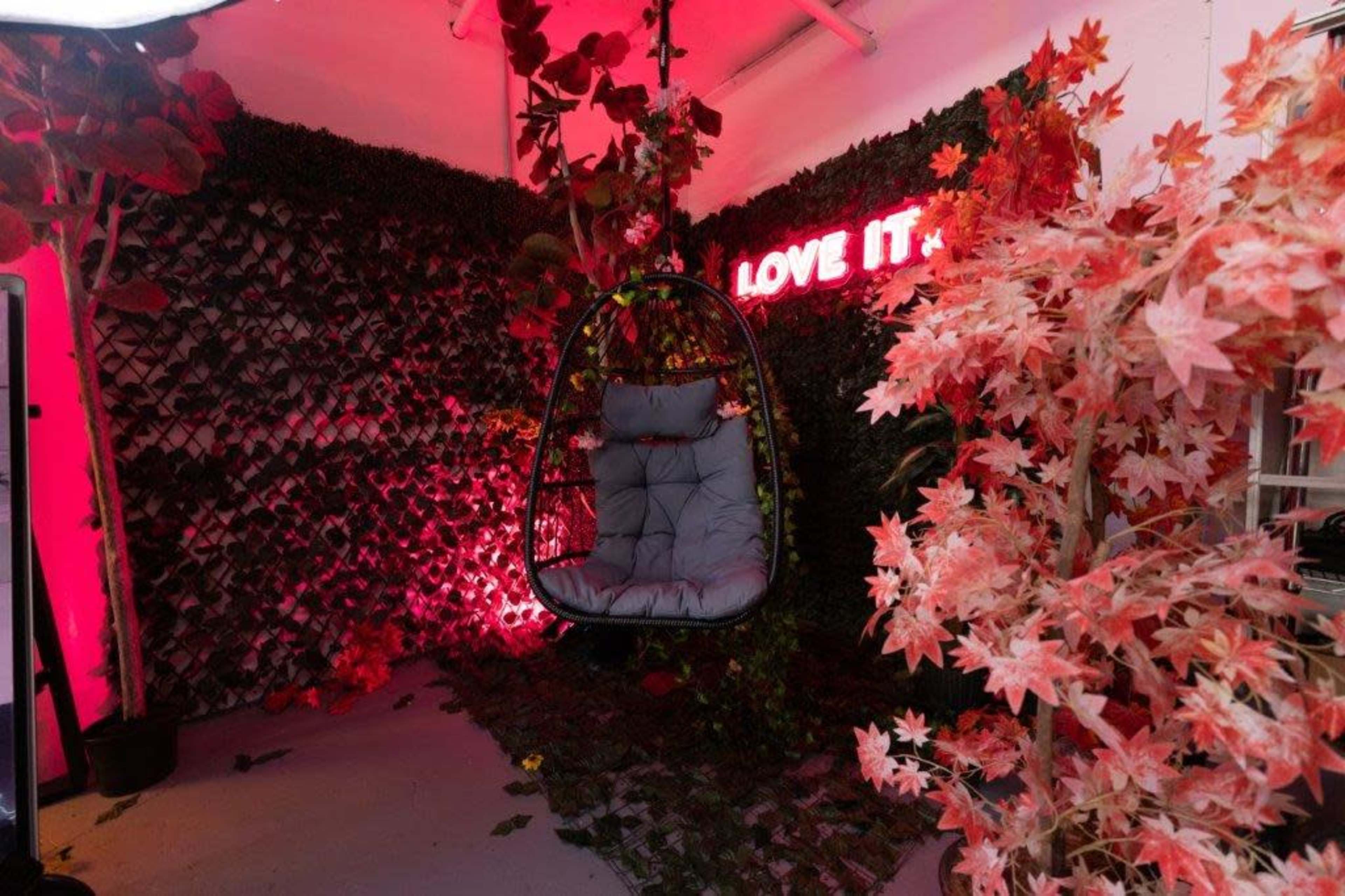 A hanging chair is positioned against a backdrop of green foliage and bright red artificial plants, with a neon sign that reads "LOVE IT" illuminating the scene.