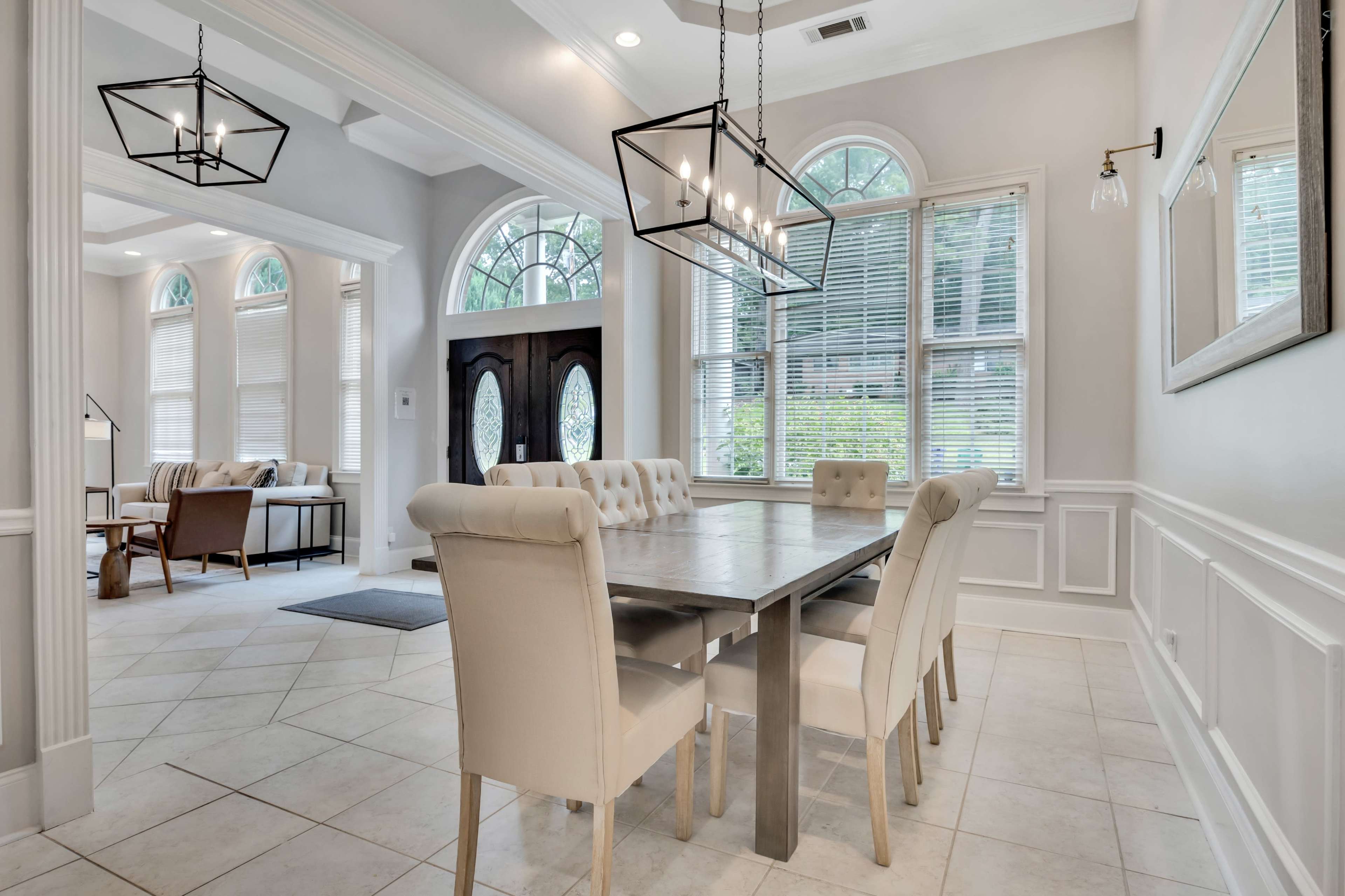 A well-lit dining area featuring a large table surrounded by cream-colored chairs, adjacent to a living space with large windows.