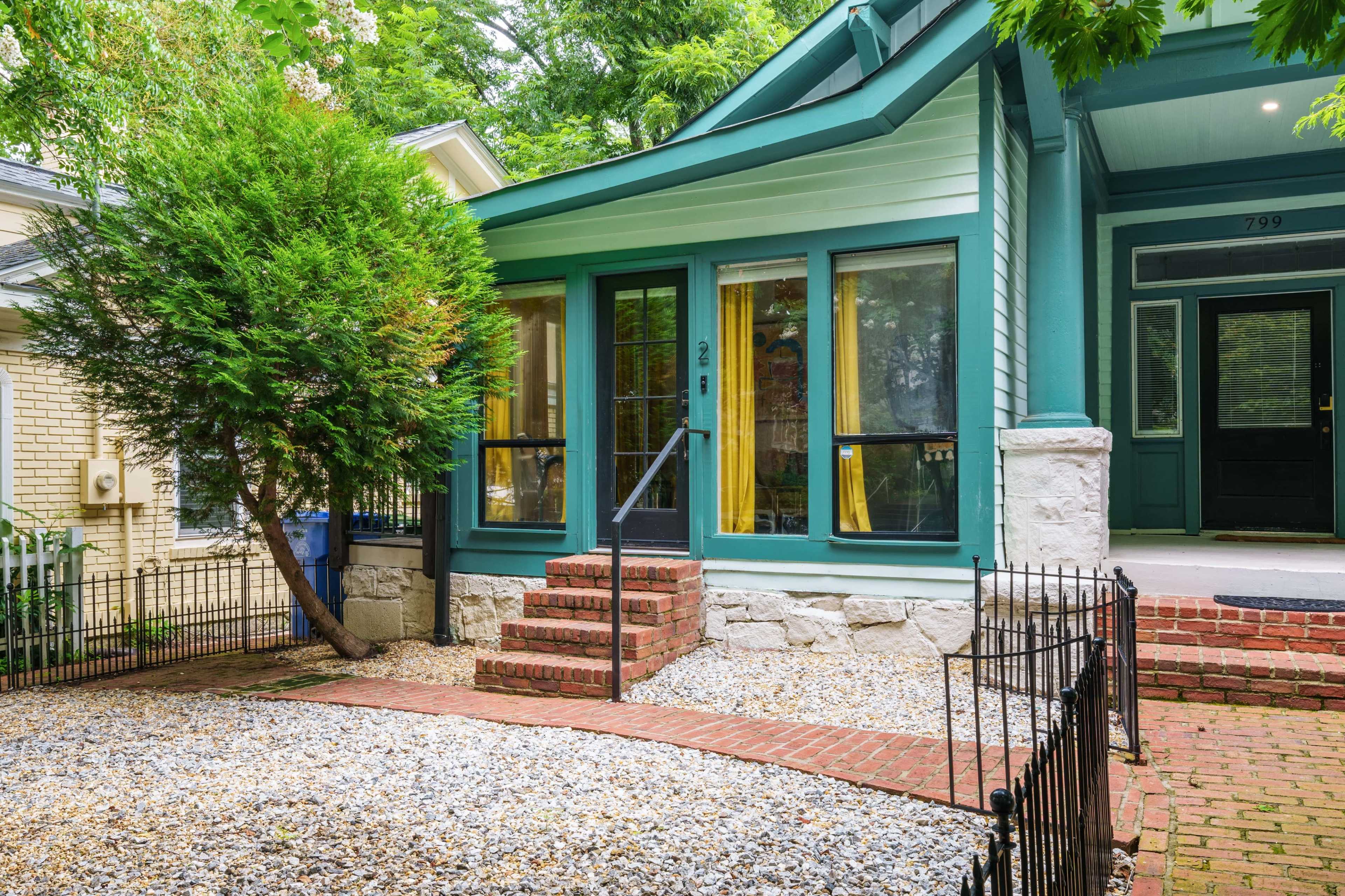The image shows a residential house with a green porch, steps leading to the entrance, and a gravel landscape bordered by a fence.