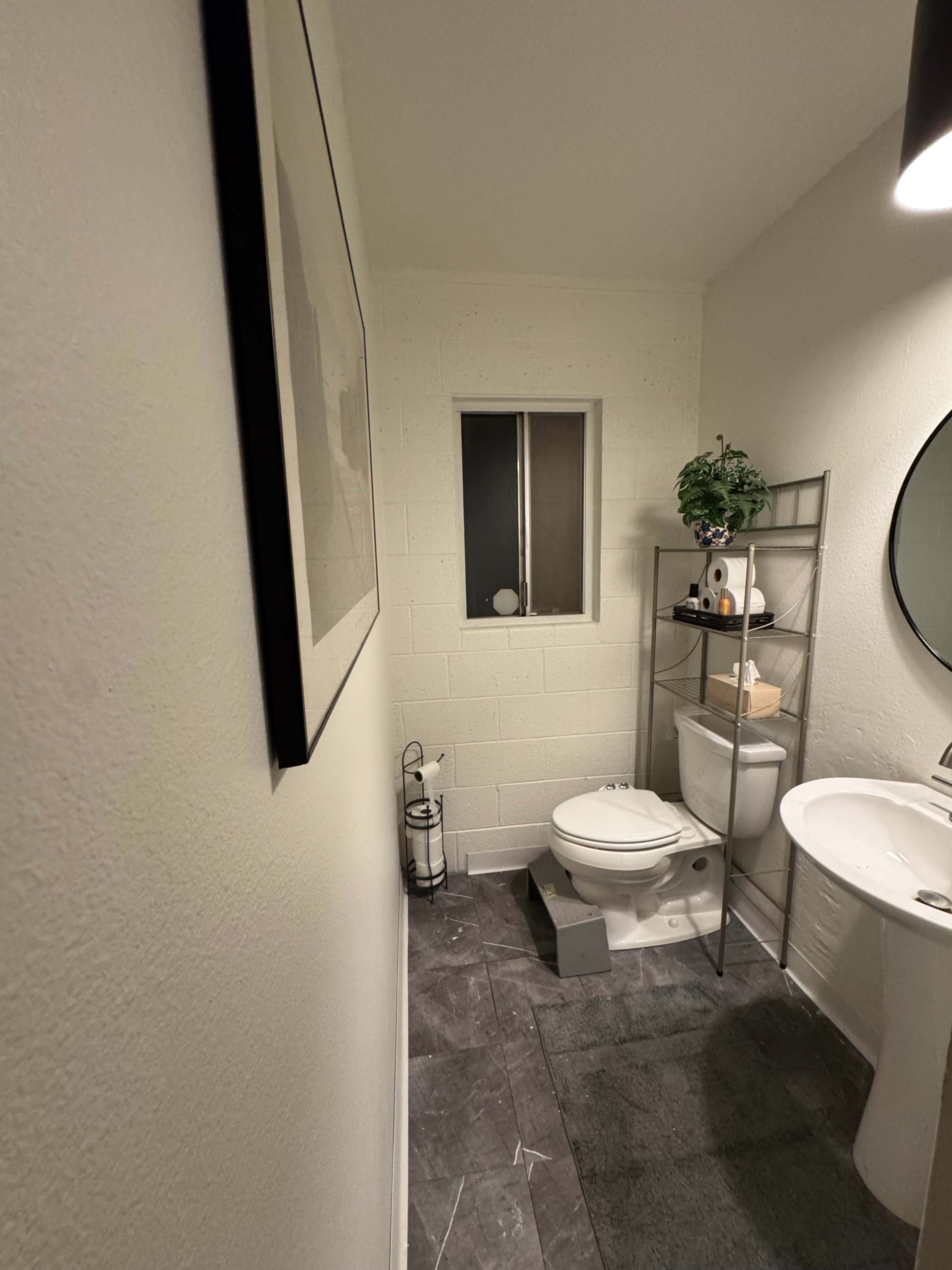 The image shows a small bathroom with a toilet, a sink, a mirror, and a shelf with decorative items.