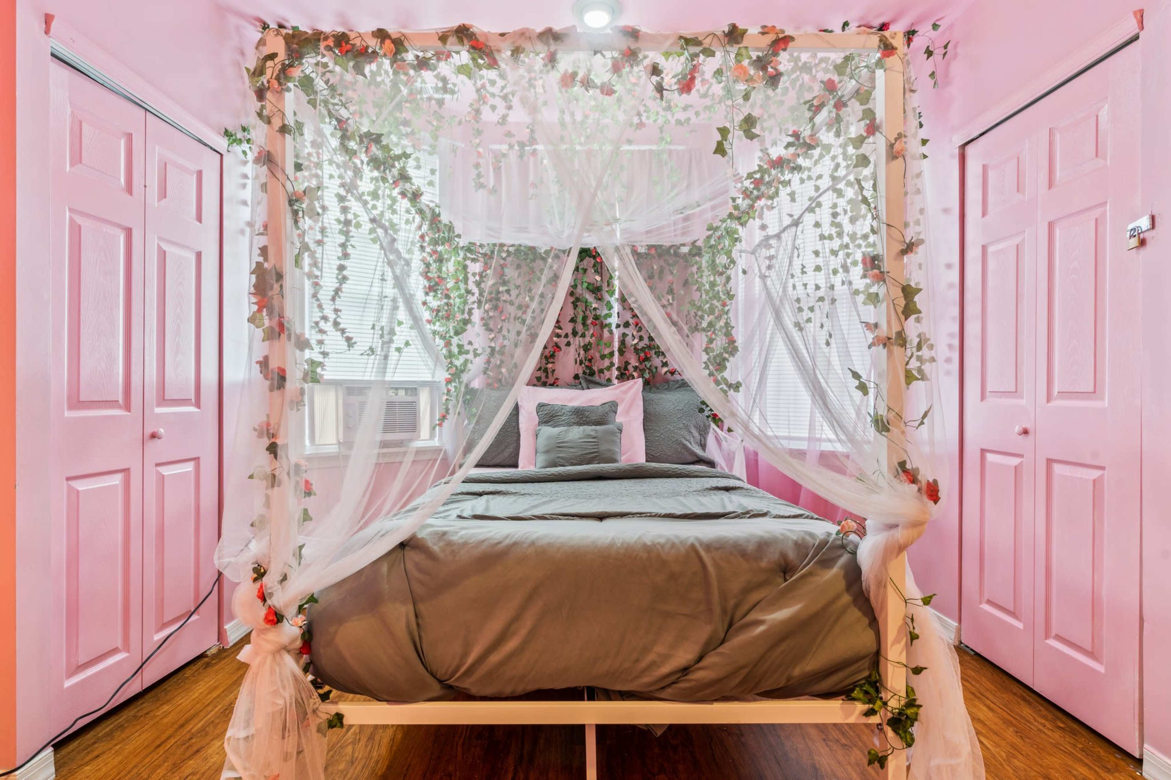 A bedroom features a canopy bed draped in sheer fabric and adorned with greenery, set against pink walls and closed doors.