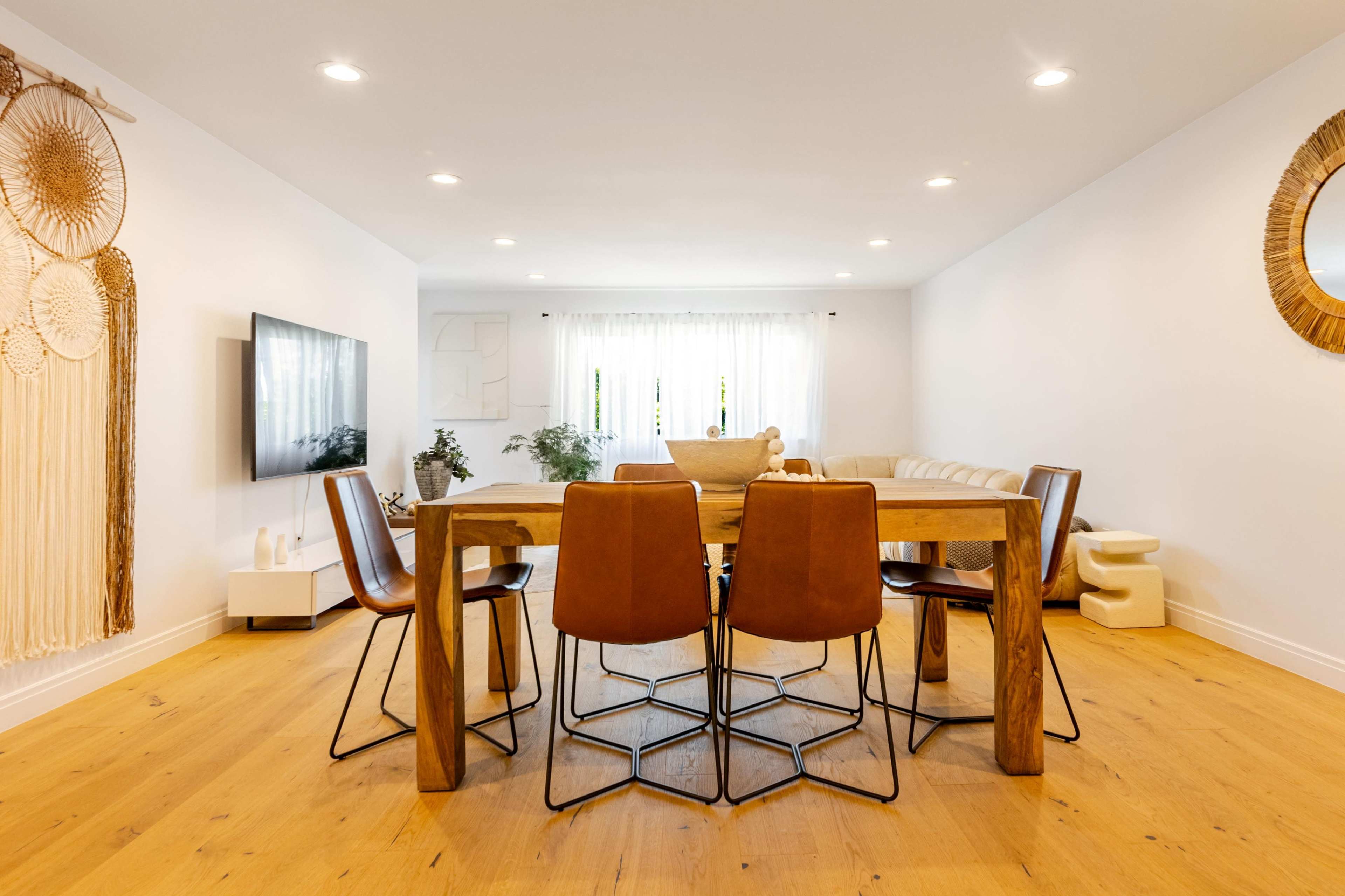 The scene depicts a modern dining area featuring a wooden table surrounded by six brown leather chairs, with a television and a decorative wall hanging in the background.