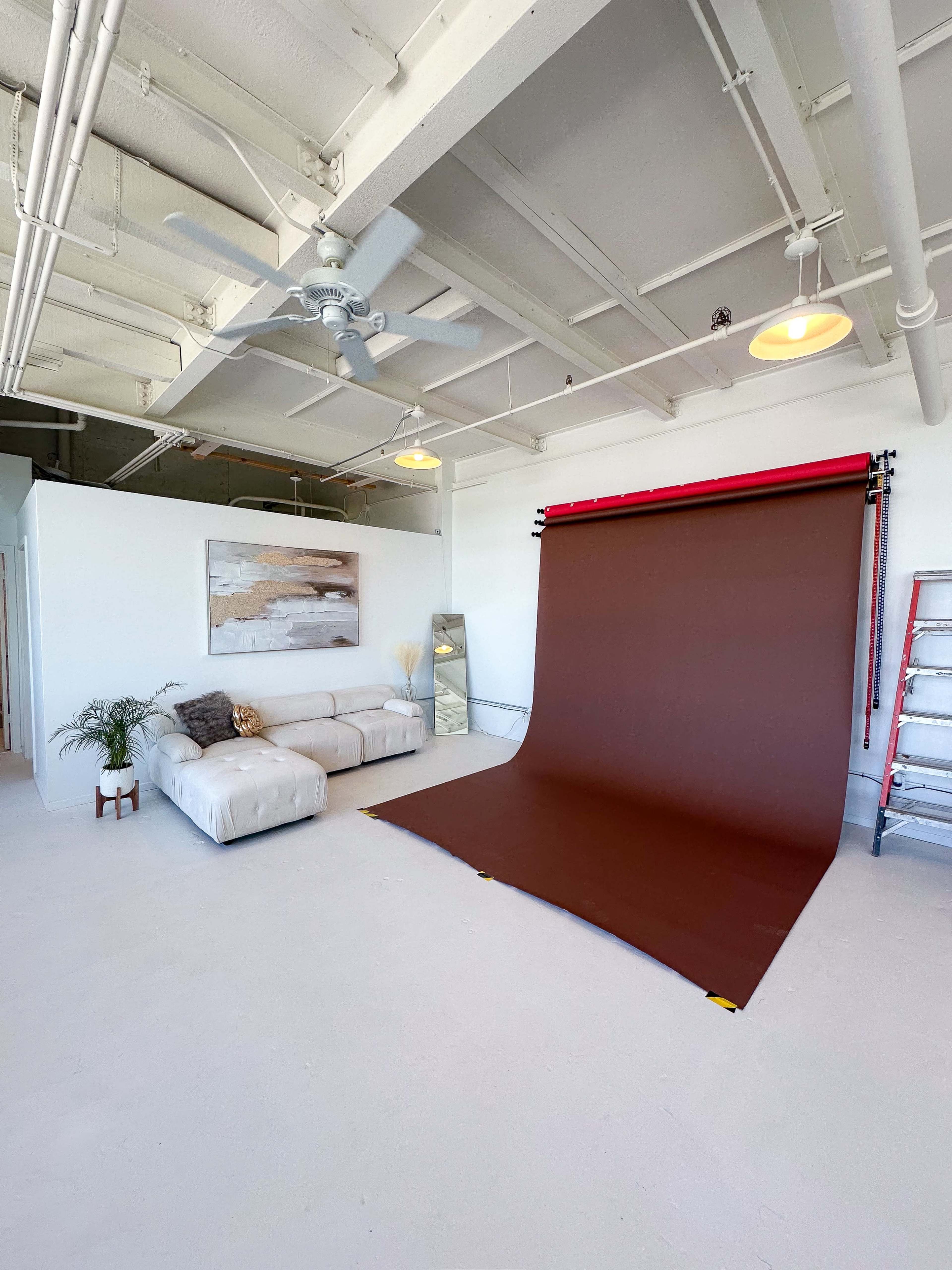 The image shows a bright studio space featuring a beige couch, a brown photography backdrop, and a ladder against the wall.