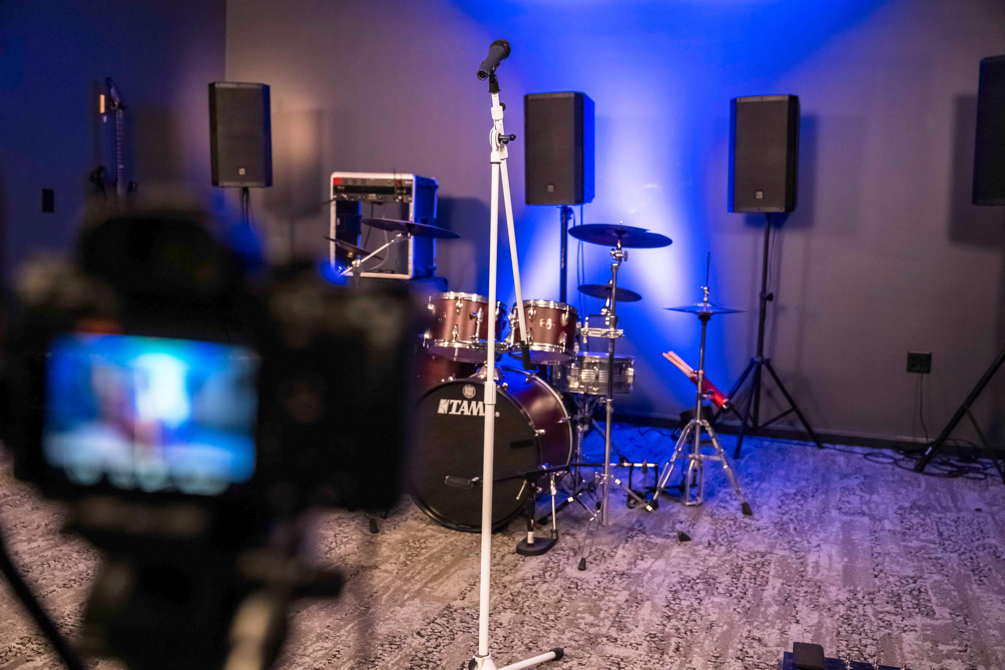 A microphone stand is positioned in front of a drum set with amplifiers and speakers on a stage illuminated by blue lights.