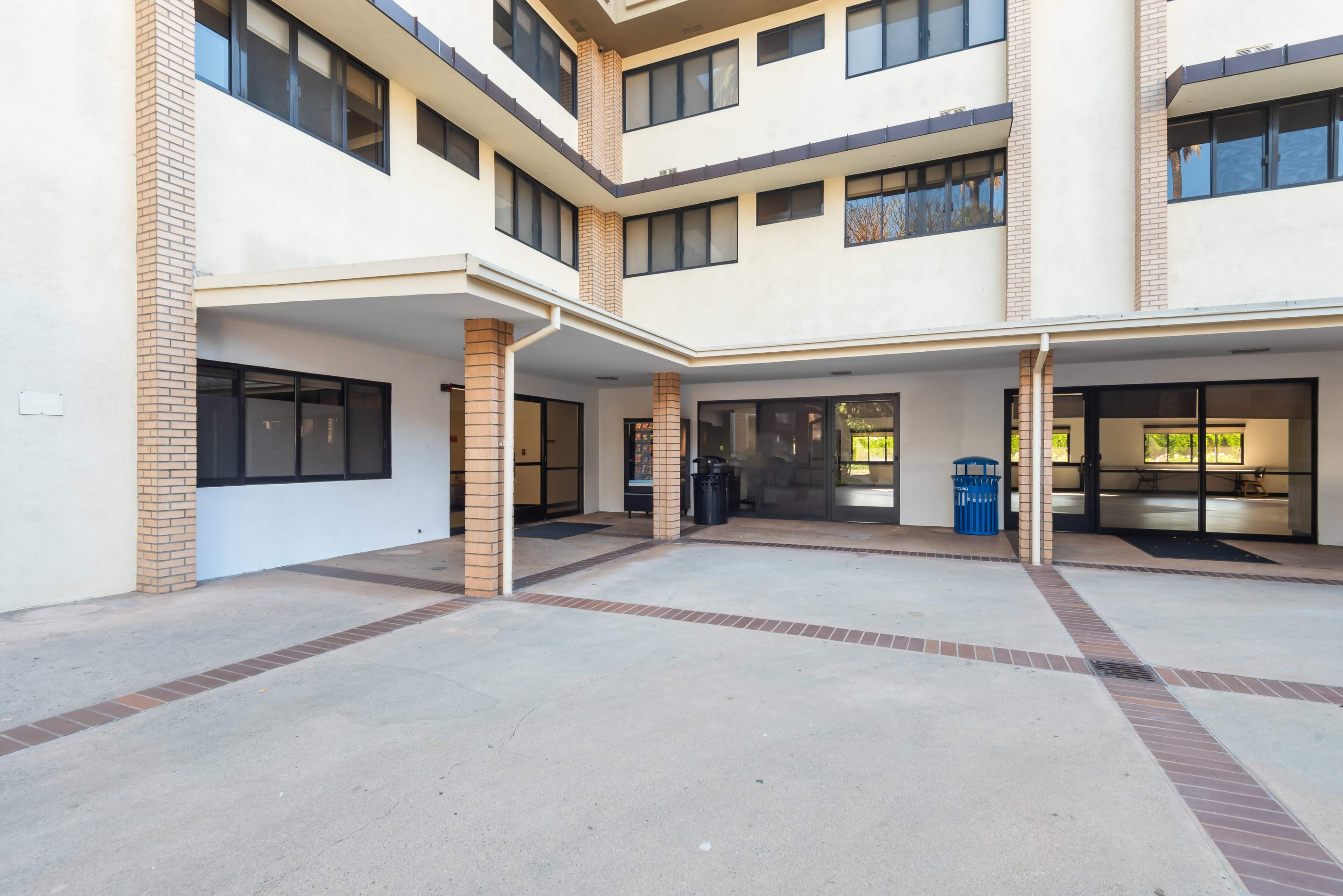 The image shows a building entrance with a paved courtyard, featuring multiple windows and a blue trash bin.