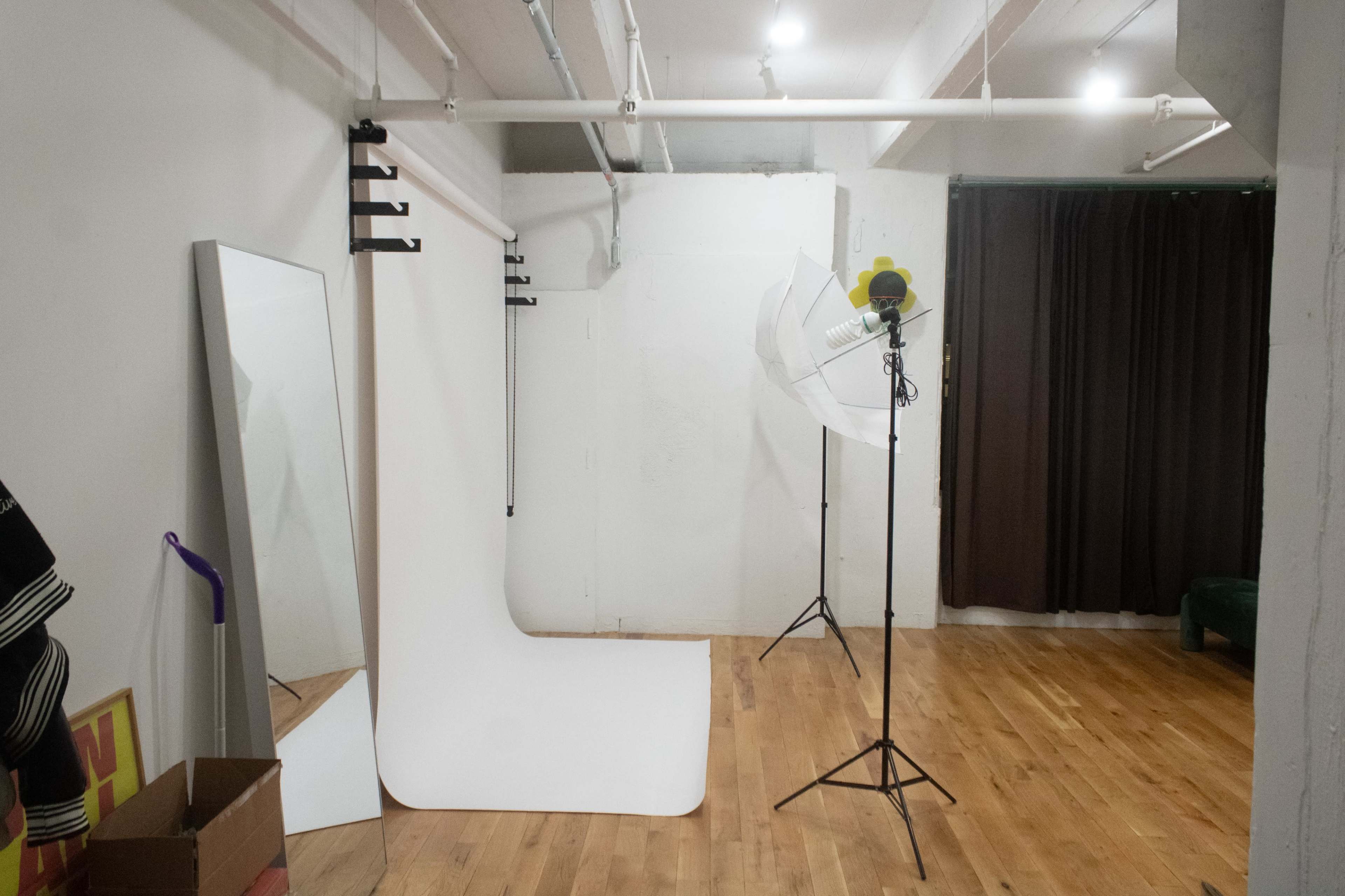 The image shows a photography studio with a white backdrop, a large light umbrella, and a mirror against a wall.