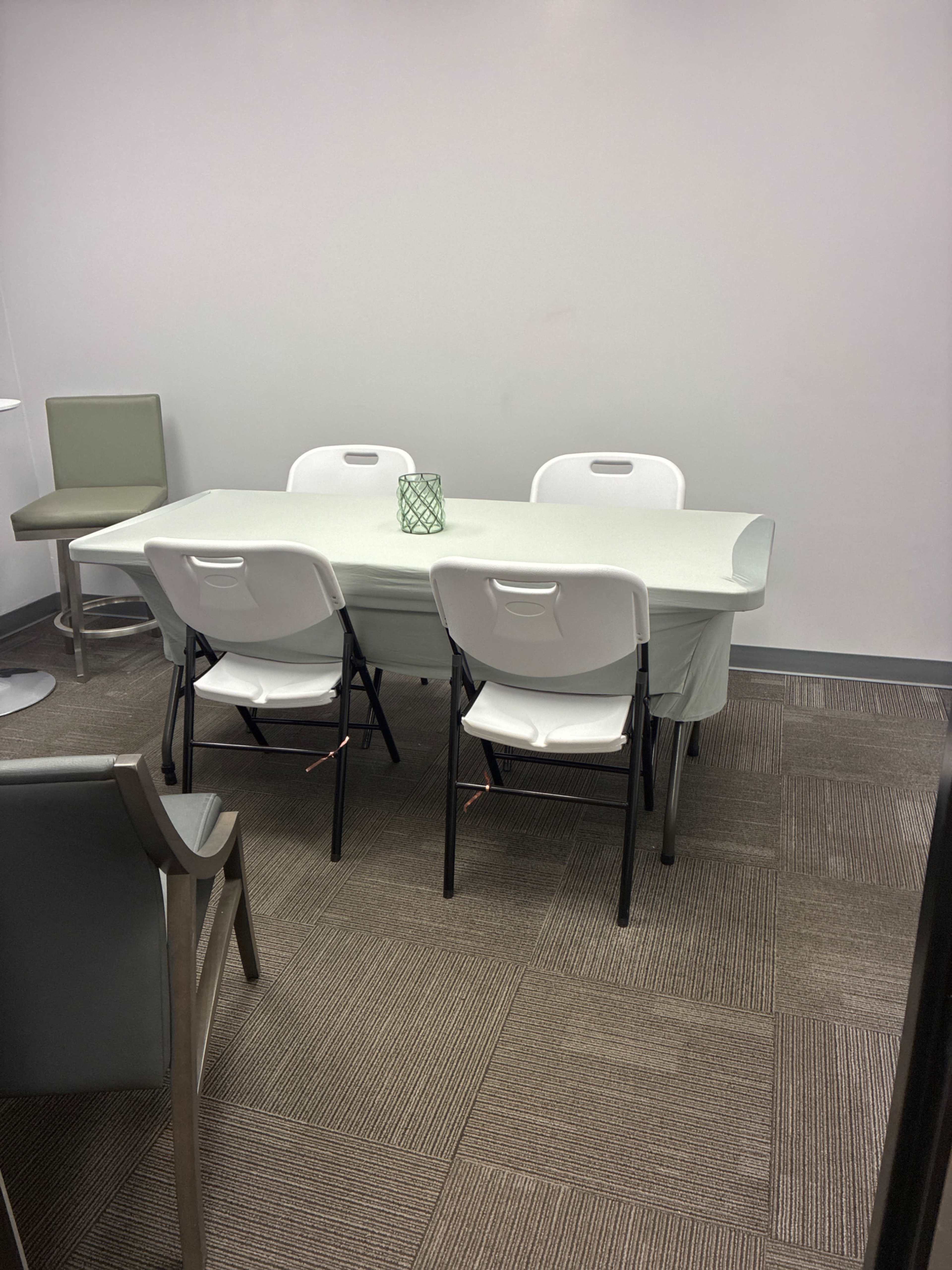 The image shows a small meeting room with a rectangular table covered in a light cloth, surrounded by four white folding chairs, and a decorative green item on the table.