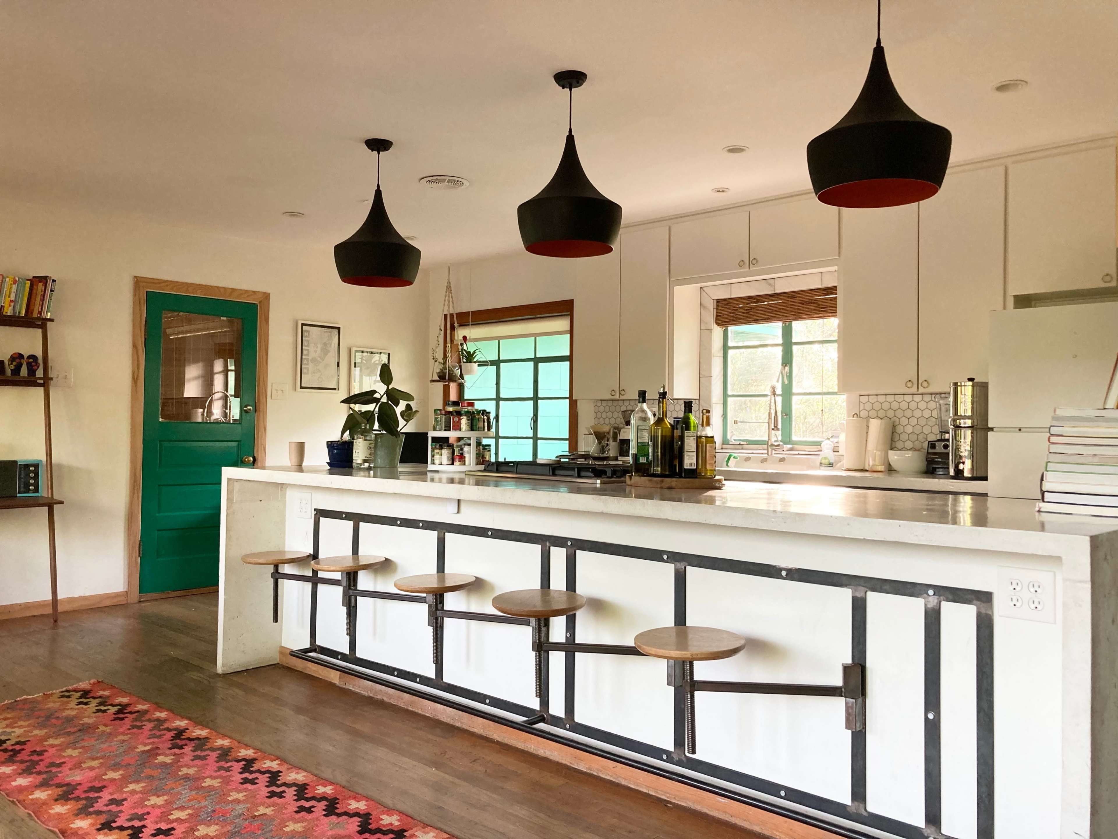 A modern kitchen features a central island with bar stools, black pendant lights, and a vibrant green door.