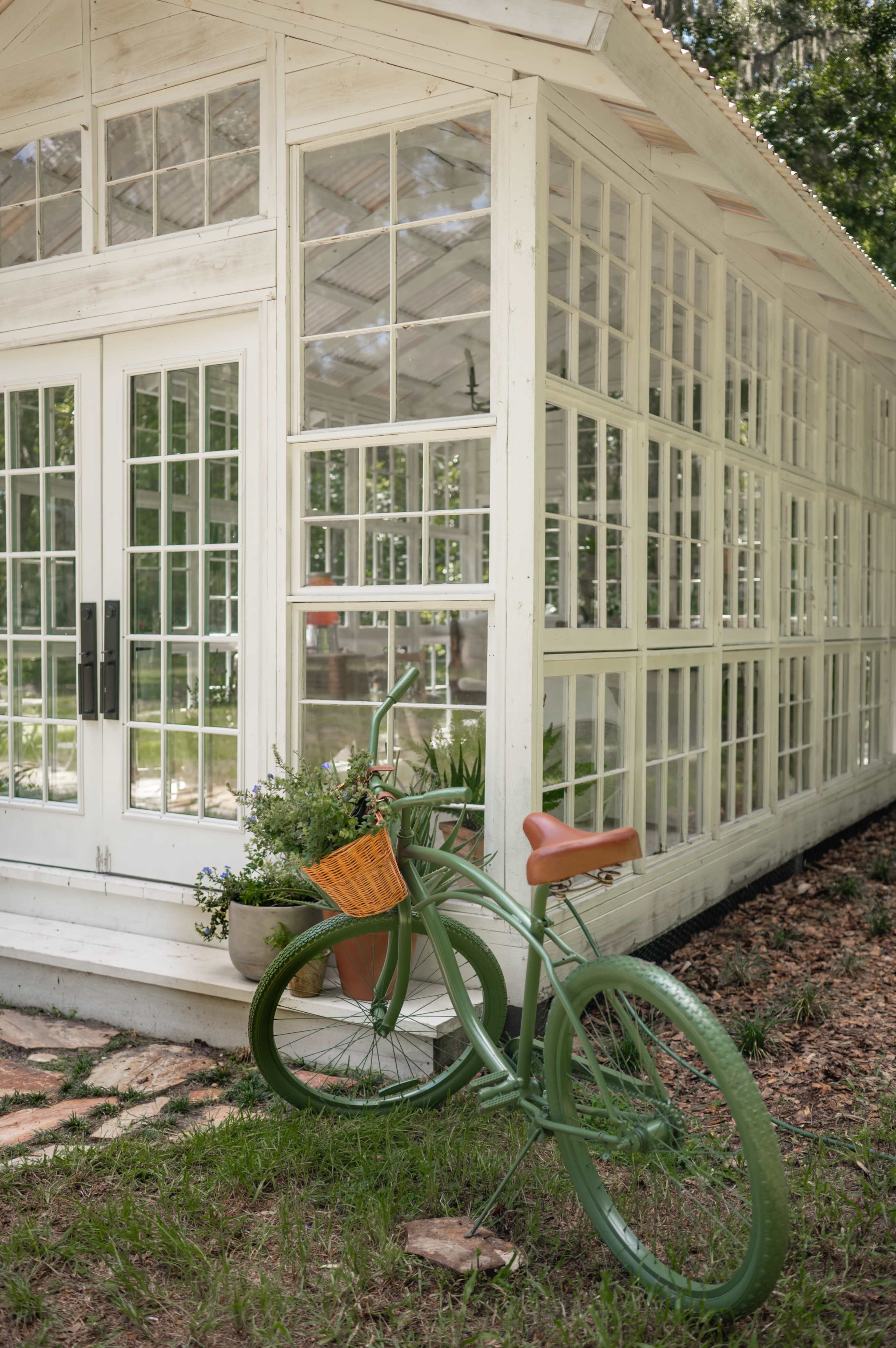 A vintage green bicycle with a basket leans against a white glass greenhouse surrounded by grass and plants.