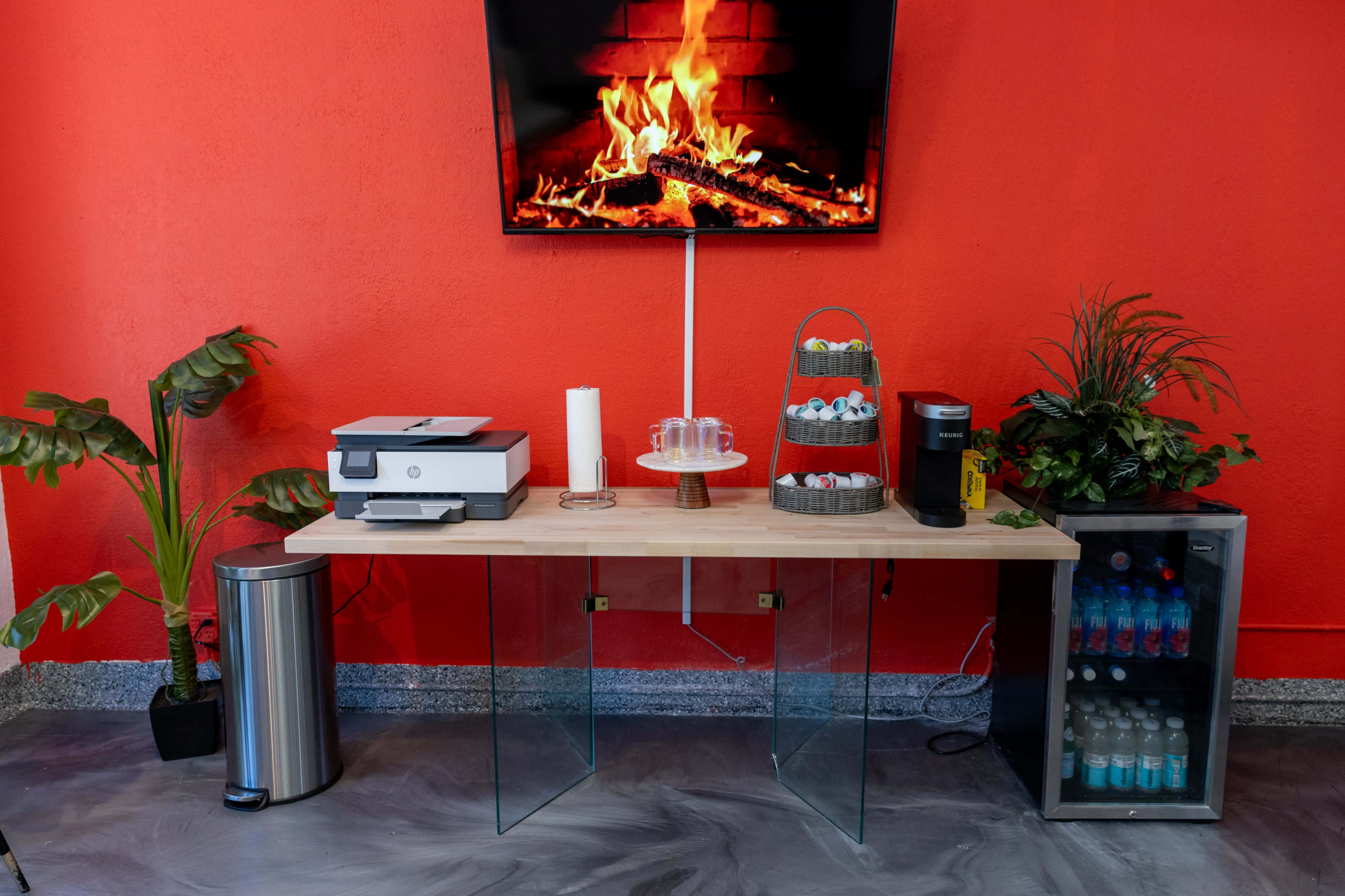 A glass-top table with a printer, snacks, and beverages is set against a red wall featuring a television displaying a fireplace.