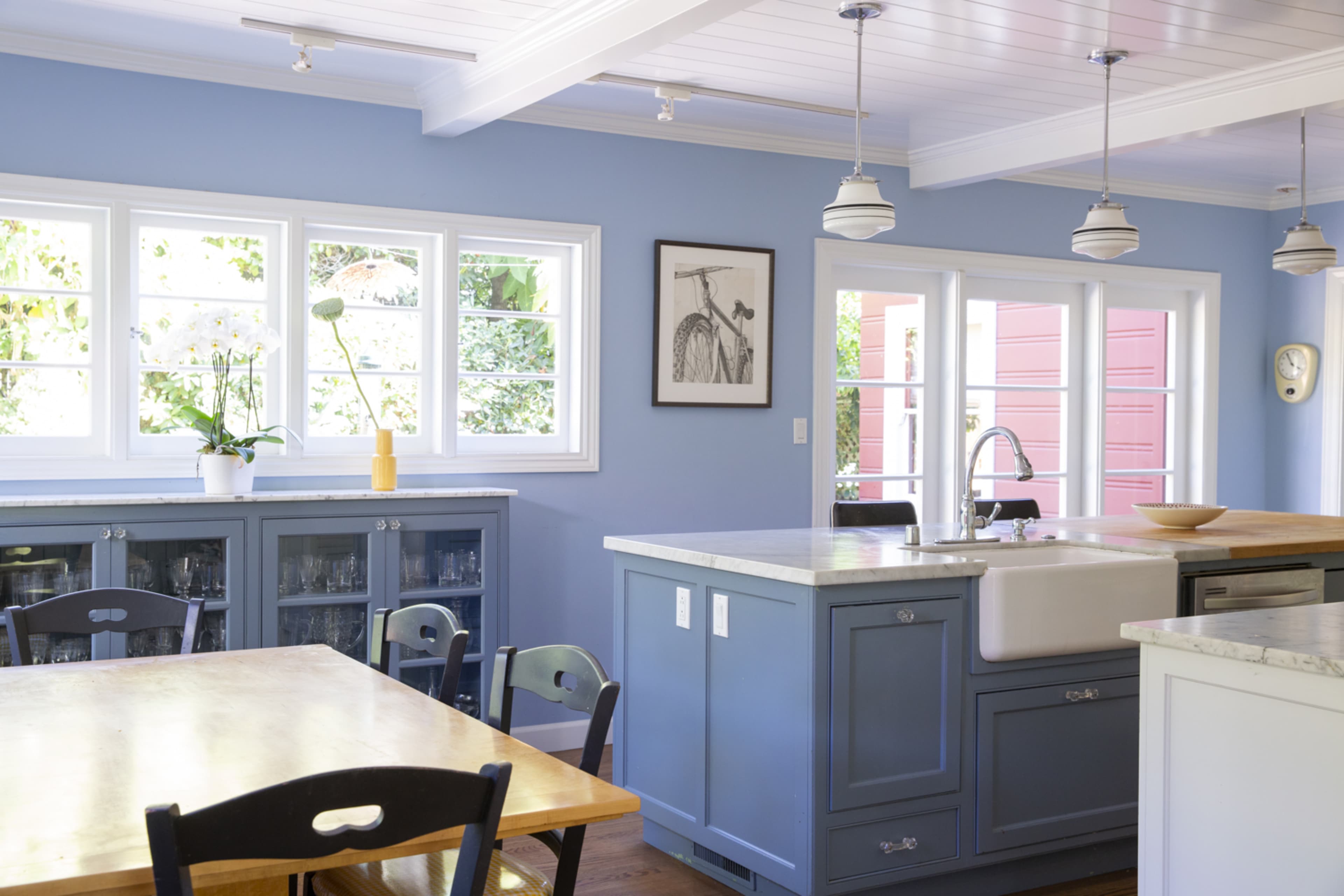 A modern kitchen features blue walls, wooden tables, and a large central island with a farmhouse sink, flanked by large windows.