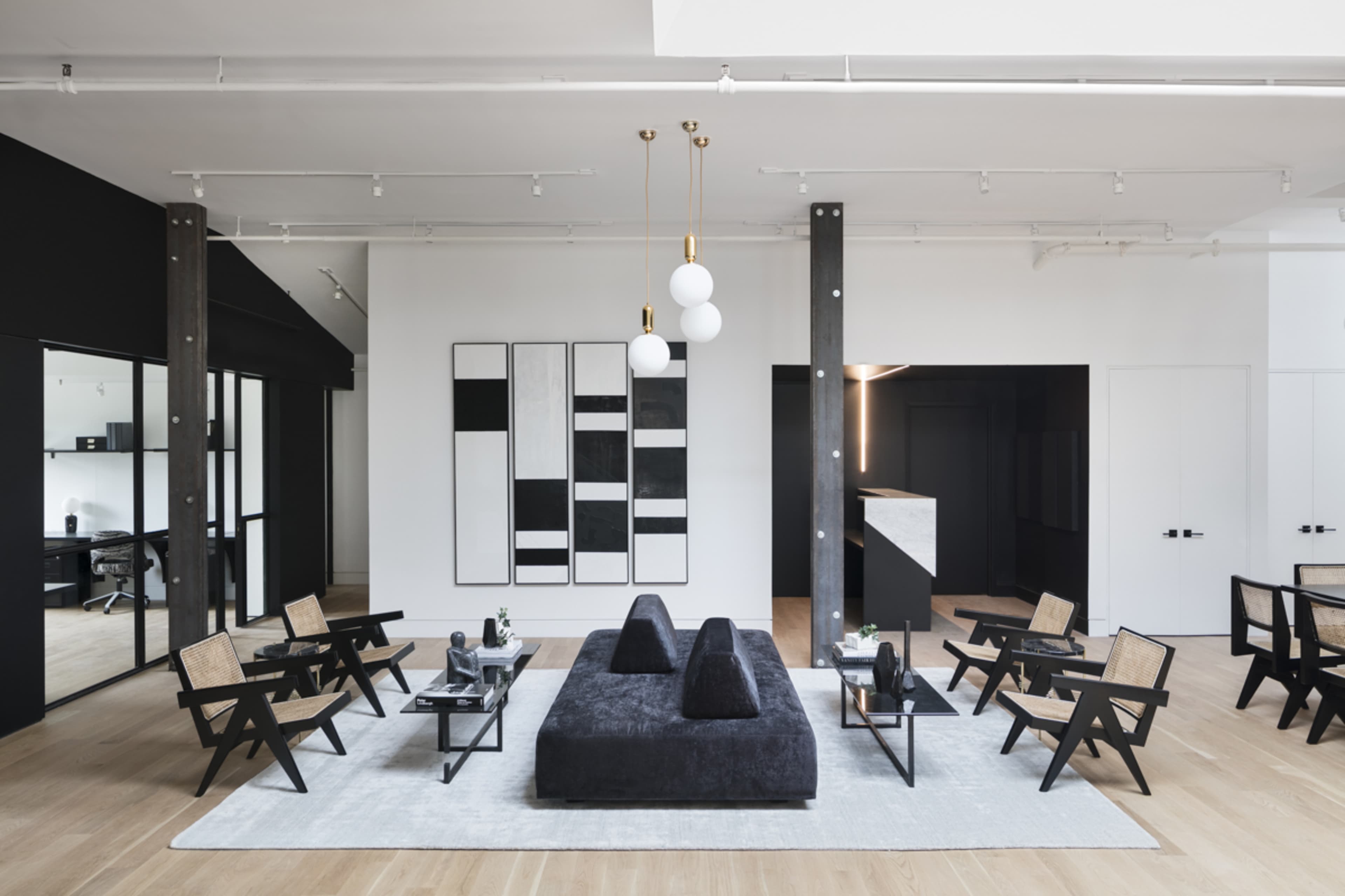 A modern interior space features a central dark sofa surrounded by unique wooden chairs and artwork on the walls, with an open layout that includes a desk area in the background.