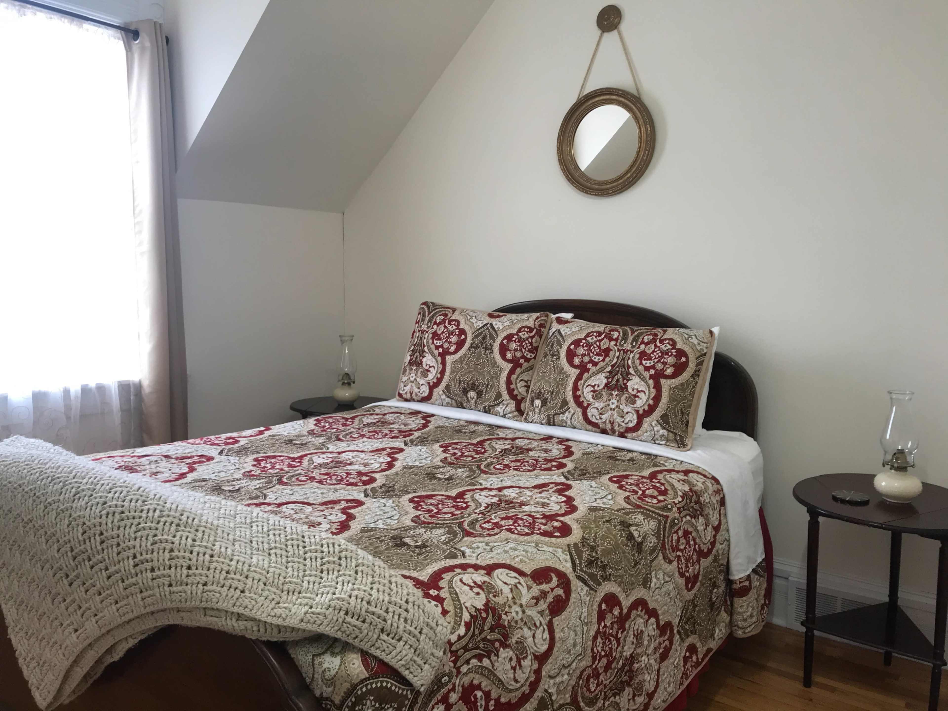 A neatly made bed with a patterned quilt and decorative pillows is positioned in a well-lit room featuring a large window and a small side table.