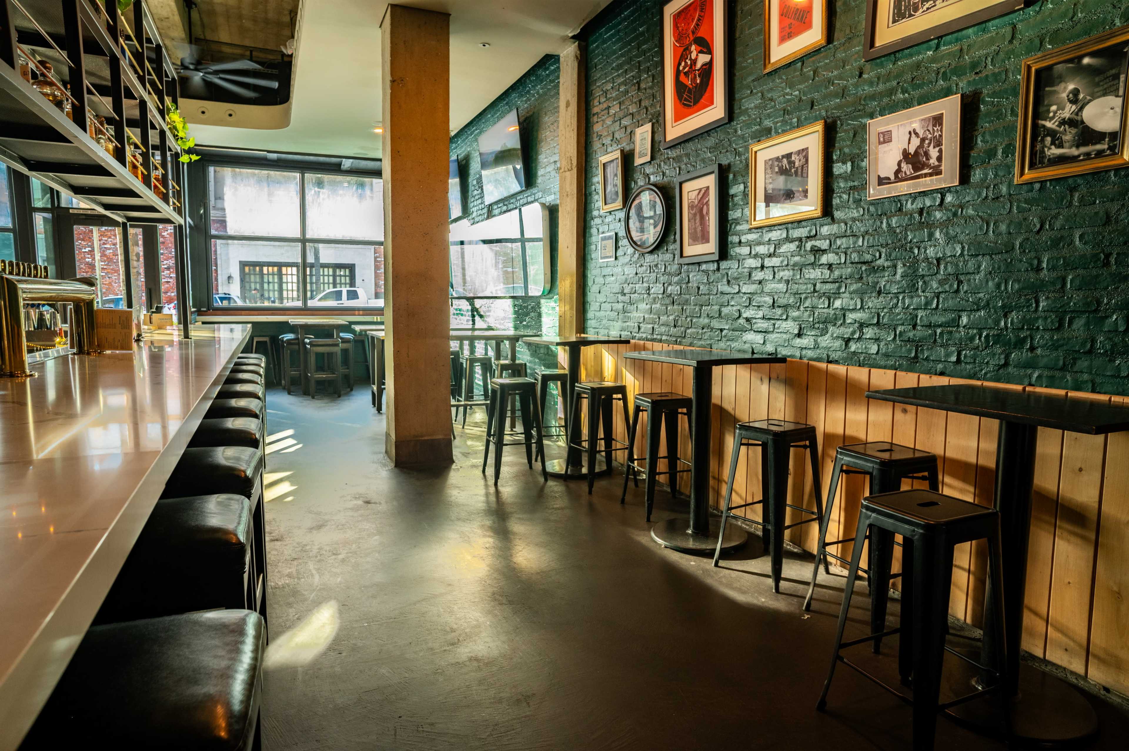 The image shows a modern bar with a long counter, high stools, and a brick wall adorned with framed photographs.