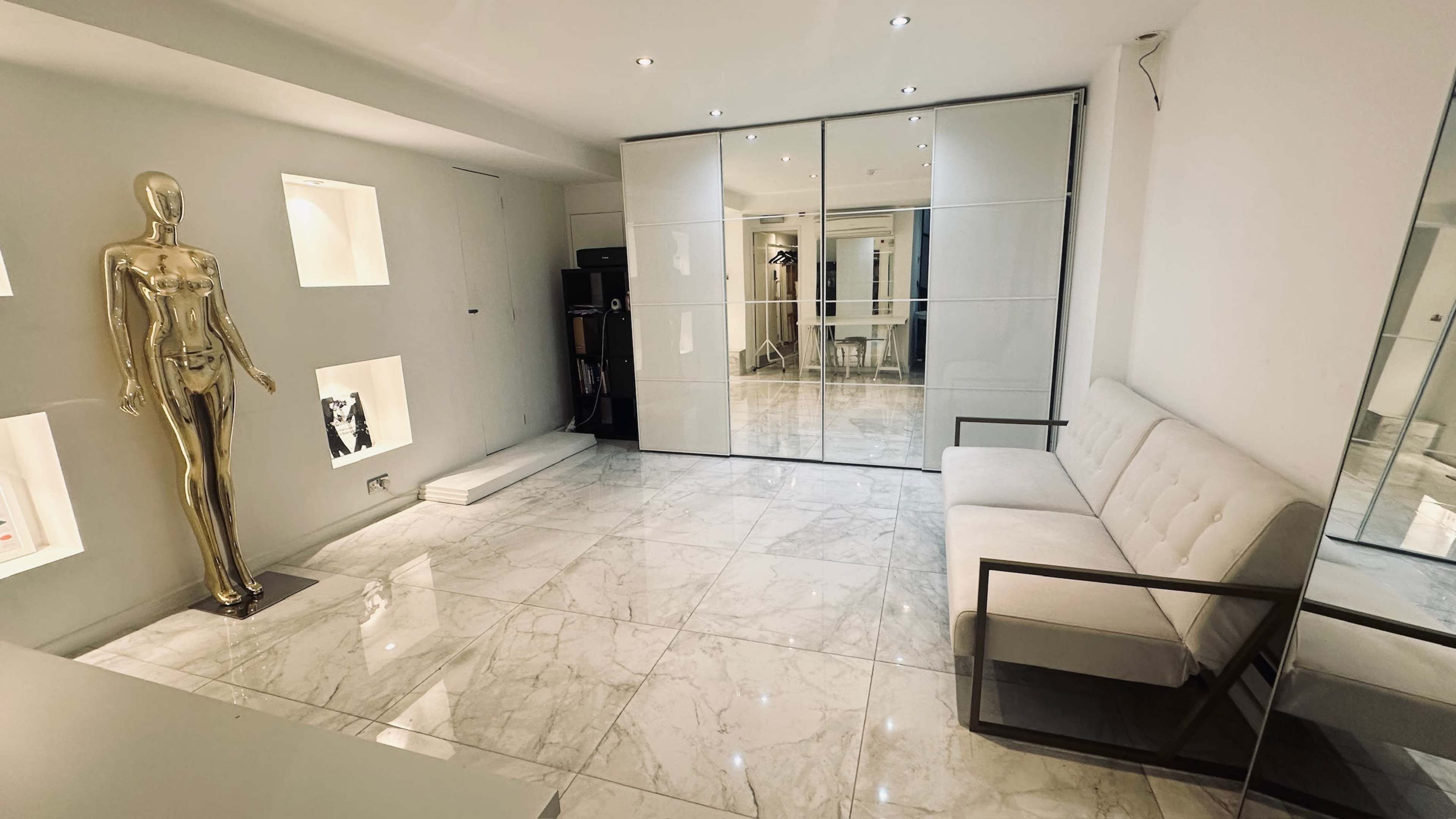 The image shows a minimalist interior space featuring a gold mannequin on the left, a white couch in the foreground, and a mirrored sliding door in the back, all situated on a glossy marble floor.