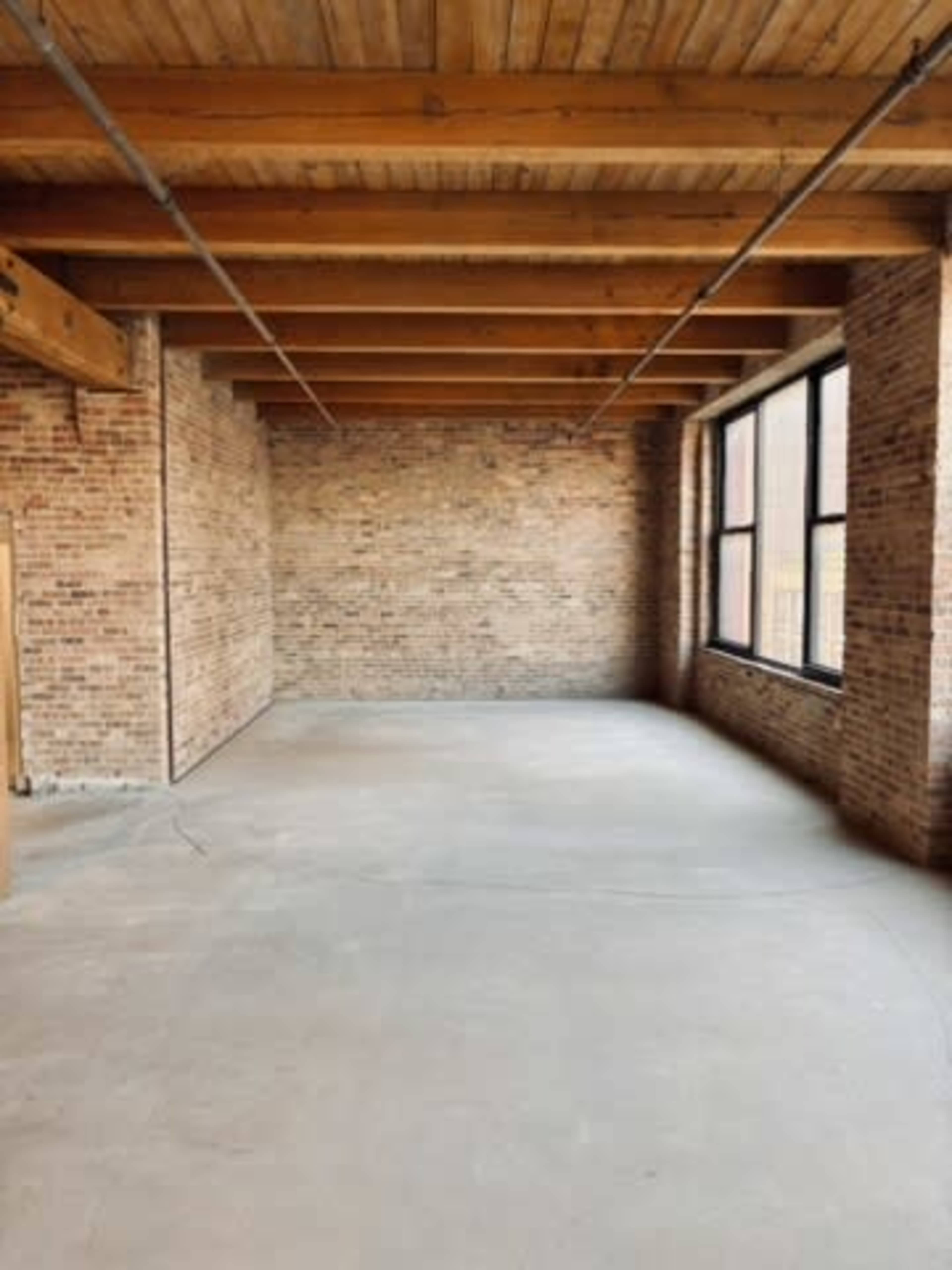 The image shows an empty room with exposed brick walls, wooden beams, and large windows.