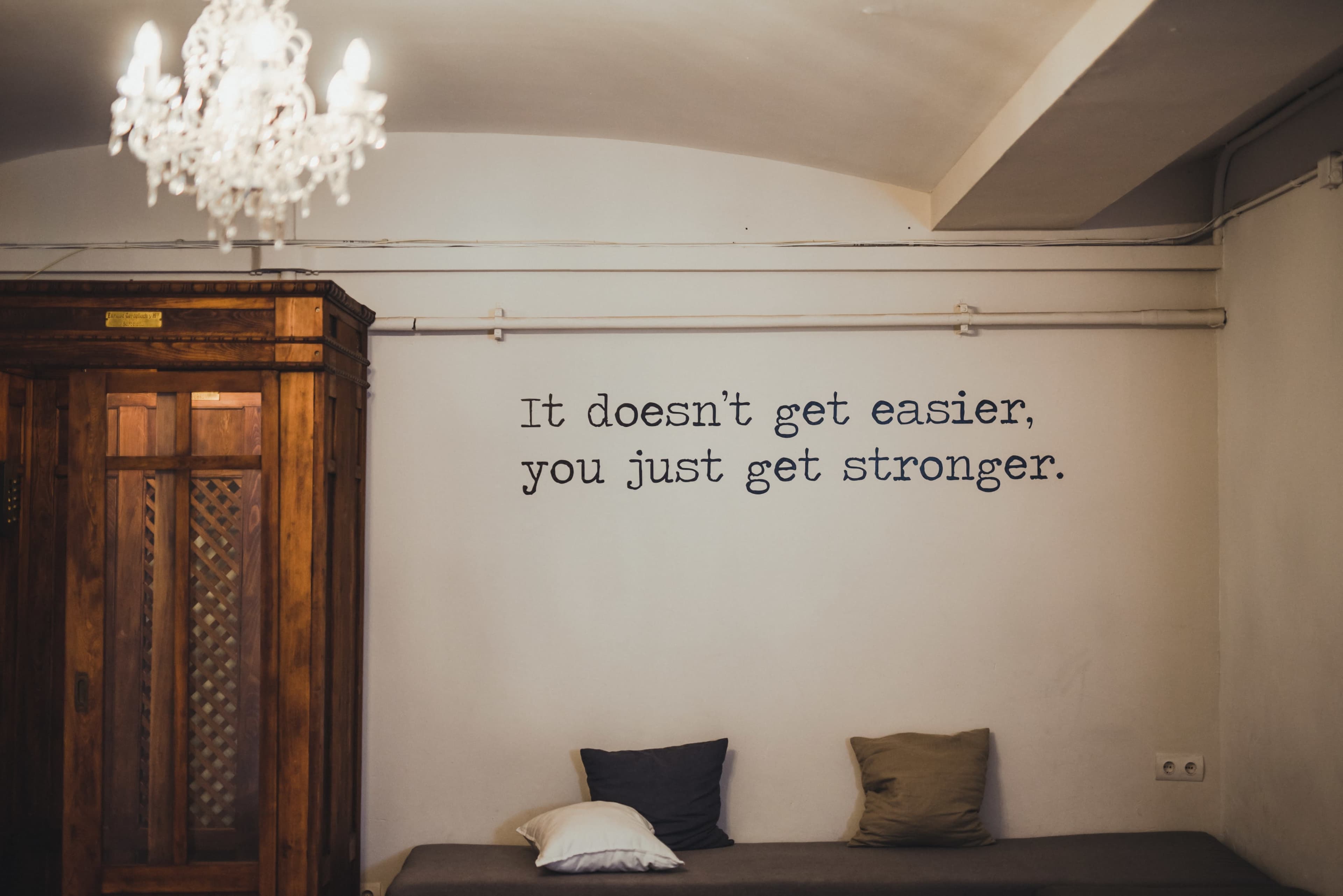 A room with a simple wall featuring a motivational quote, a wooden cabinet, and a sofa with two cushions.
