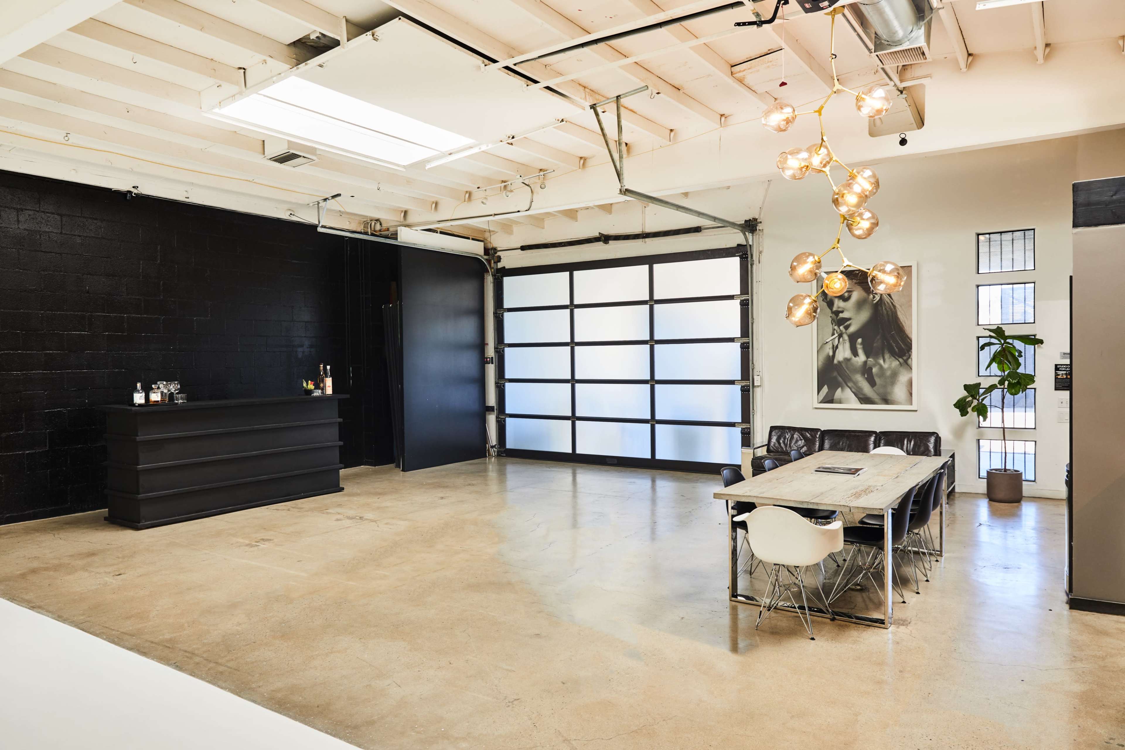The image shows a spacious, modern office with a large wooden table, a black bar counter, and a distinctive light fixture hanging from the ceiling.